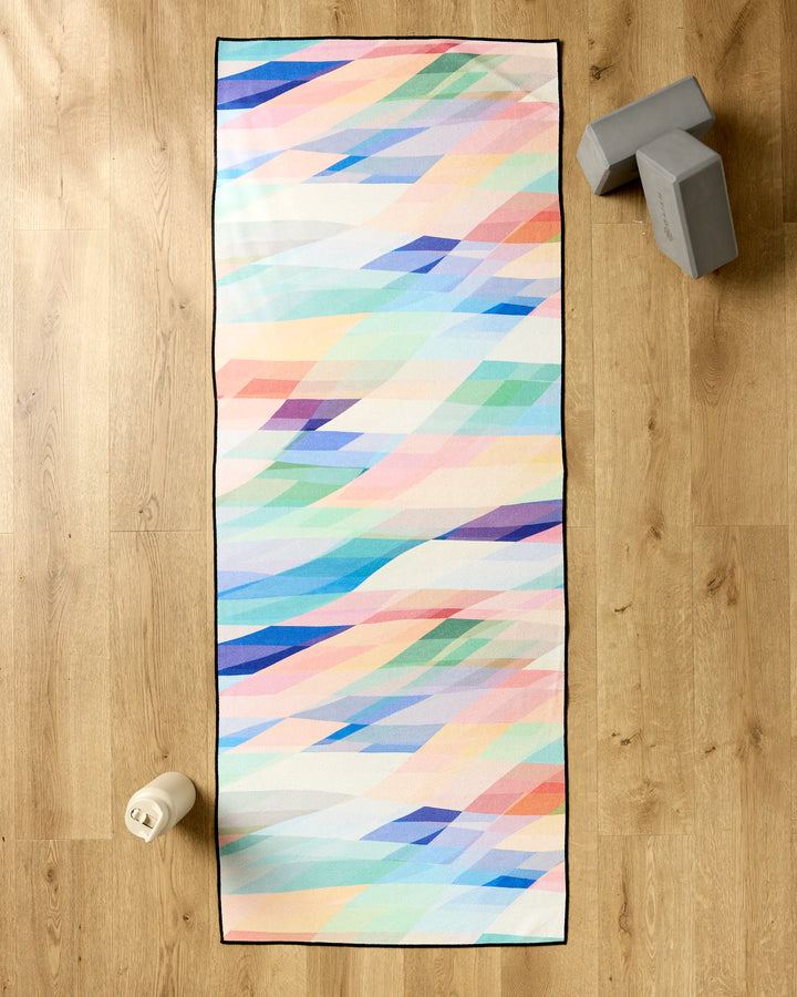 The Rachel Parker Eventide yoga mat, featuring a pastel geometric design, is spread on a wooden floor with a slip-resistant towel. Gray yoga blocks and a white water bottle are placed next to the mat.