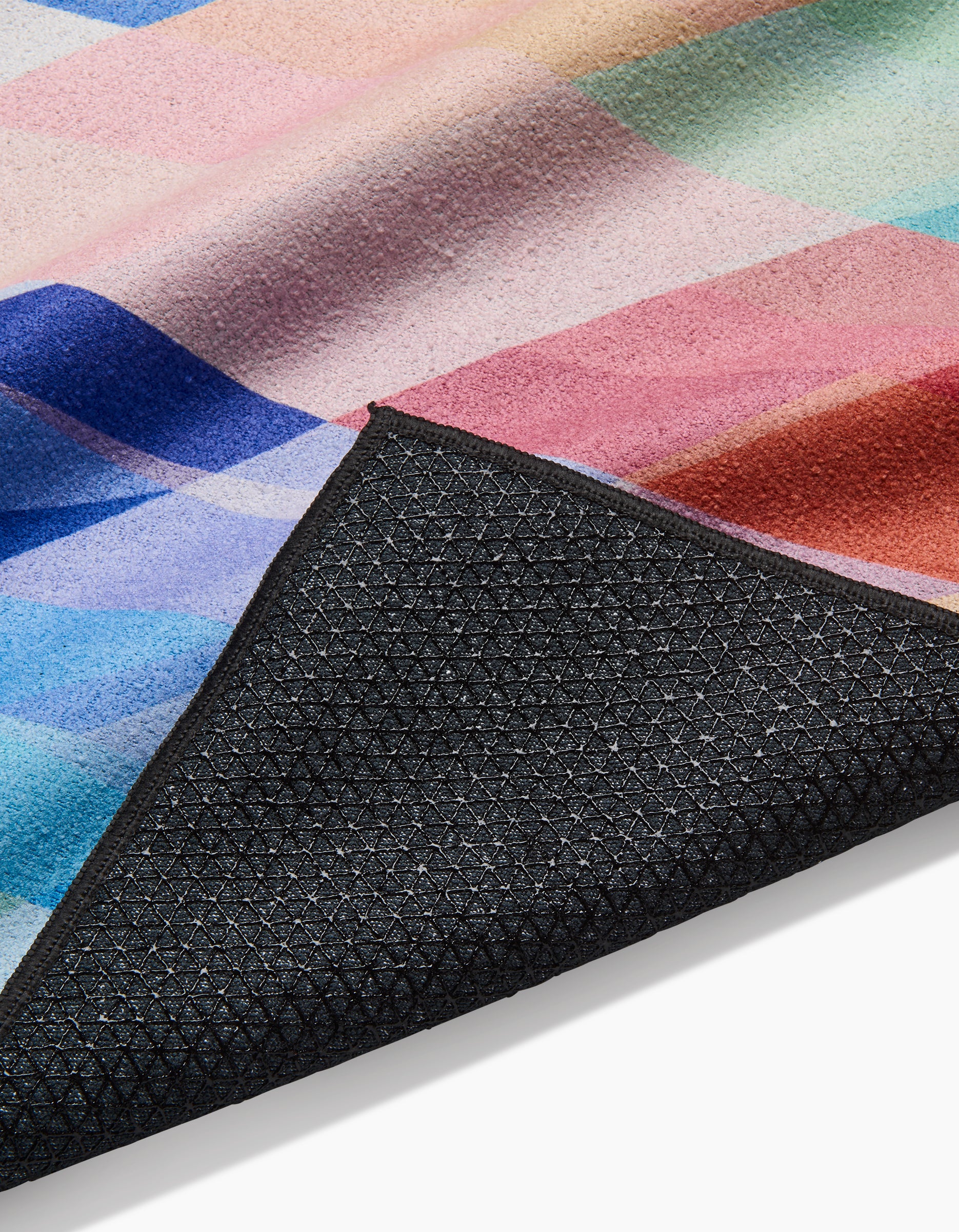 A close-up of Rachel Parkers Eventide mat shows its vibrant geometric pastel surface, with the black textured underside folded to reveal the non-slip silicone grip backing.