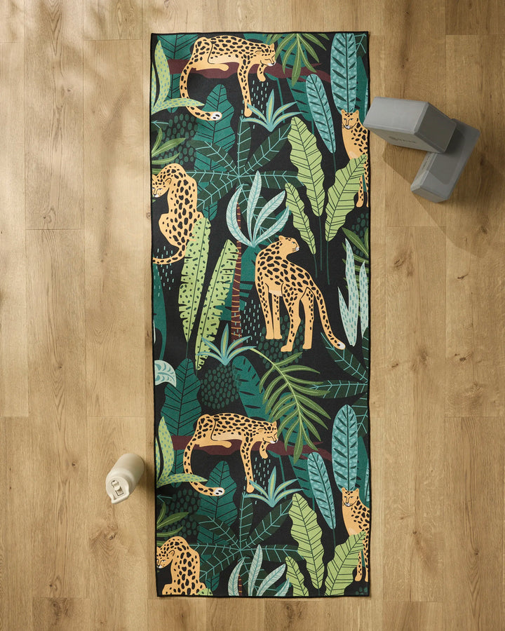 The GEOMETRY Feeling Spotty yoga mat, featuring a vibrant leopard and tropical leaf pattern, is laid on a wooden floor. Nearby are a water bottle, two gray blocks, and an eco-friendly matching towel made from recycled materials.