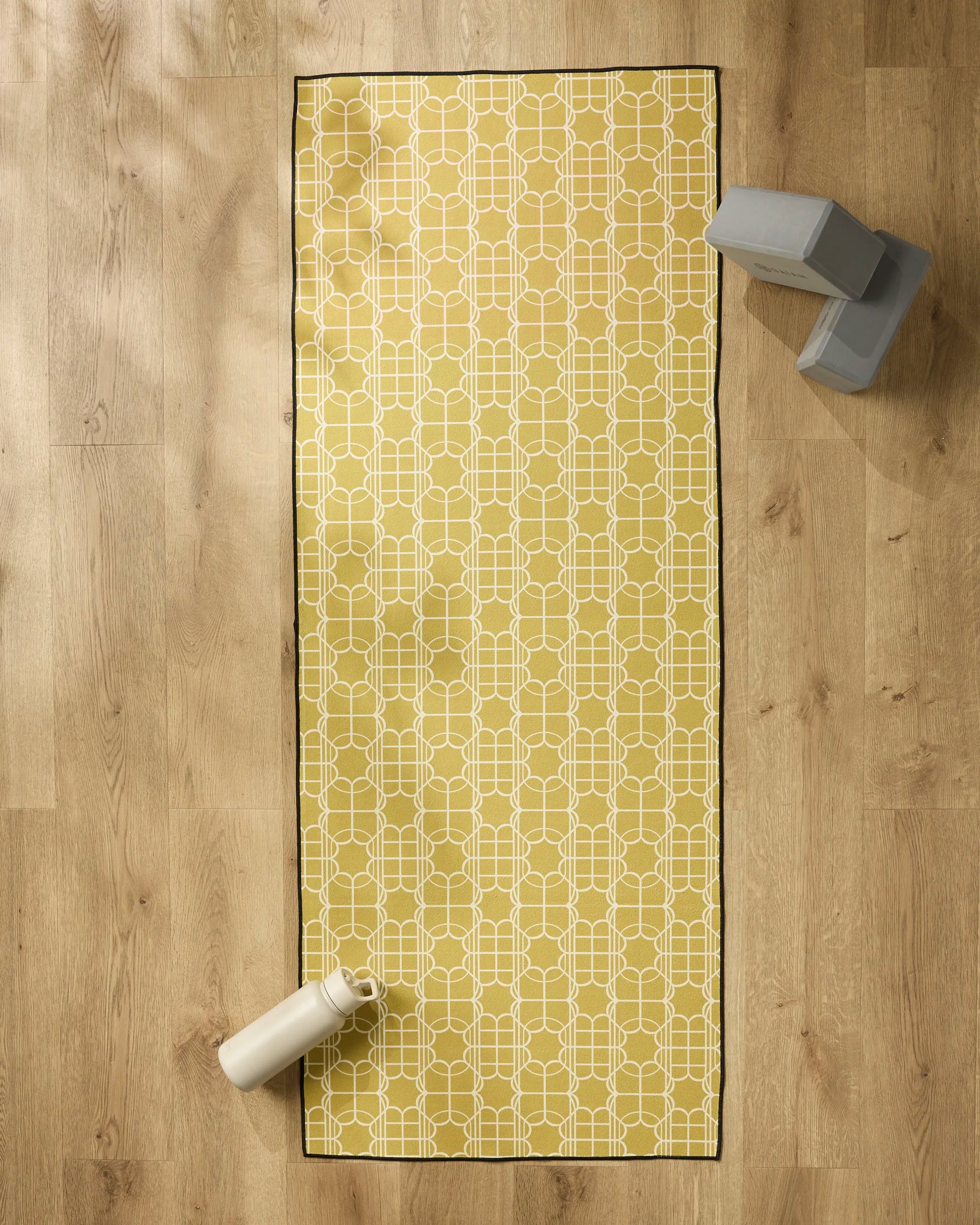 The Geometry Ferran yellow yoga mat with a white geometric pattern is on a wooden floor, topped with a silicone grip towel. A water bottle sits at one end and two gray yoga blocks are stacked in the top right corner.
