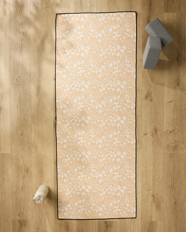 The Hufton Studio Floating Meadow yoga mat with a white floral design lies on a wooden floor. To the left is a white water bottle; at the top right, two gray yoga blocks are stacked with a non-slip towel beside them.