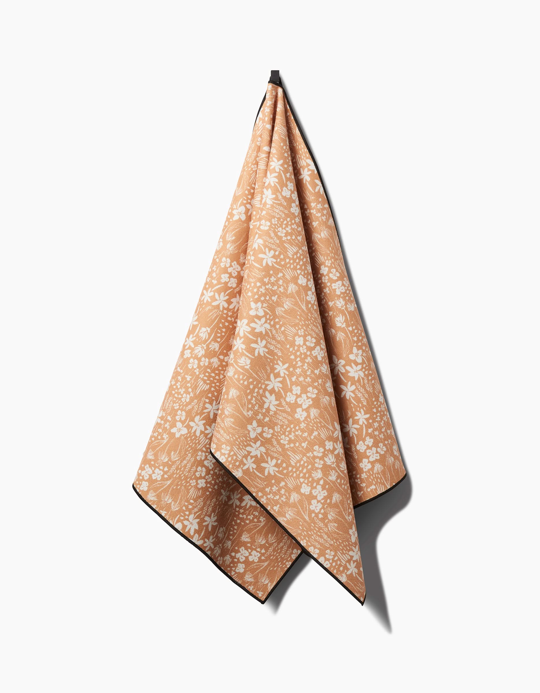 The Floating Meadow yoga towel by Hufton Studio features a peach color with a white floral pattern and dark edging. Soft and drapey, it hangs from a hook against a white background, making it a stylish, slip-resistant choice for your yoga practice.