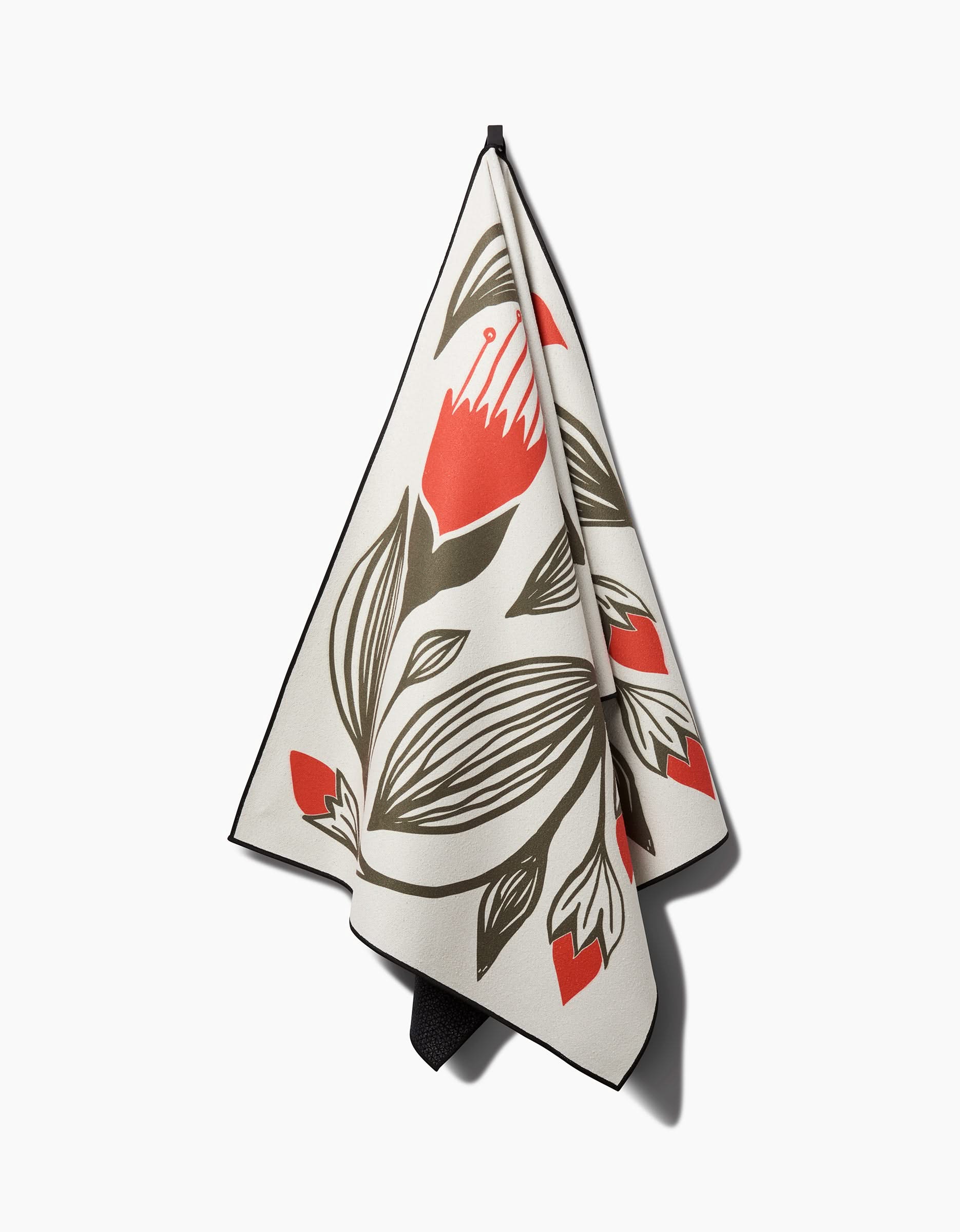The Ash Cascade Flora Stamp yoga towel, beige with bold red, black, and olive green floral patterns and silicone grip dots, hangs from a hook against a white background.
