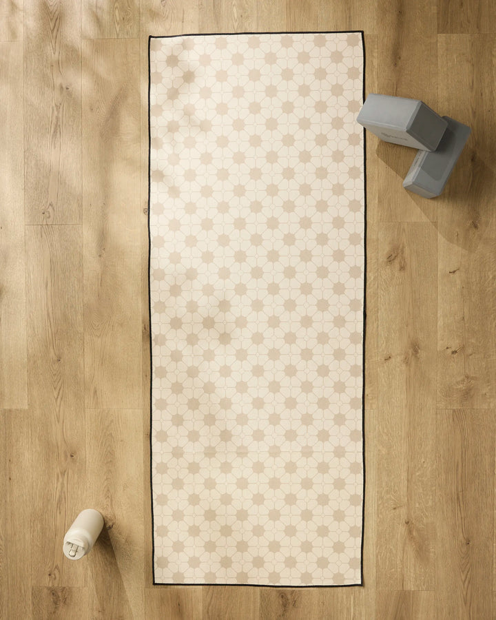 The cream-colored "Flora Tile" yoga mat by Geometry, featuring a subtle geometric pattern, lies on a wooden floor with a white water bottle at one end and two gray yoga blocks plus a slip-resistant yoga towel at the other.