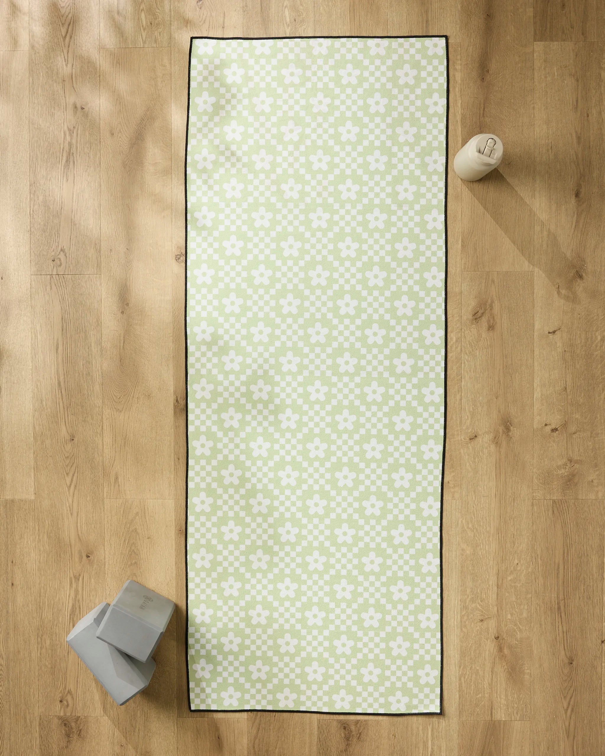 The GEOMETRY Floral Check green yoga mat with a white floral pattern lies on a wooden floor, paired with two gray yoga blocks, a white water bottle, and a slip-resistant towel for added support during your practice.