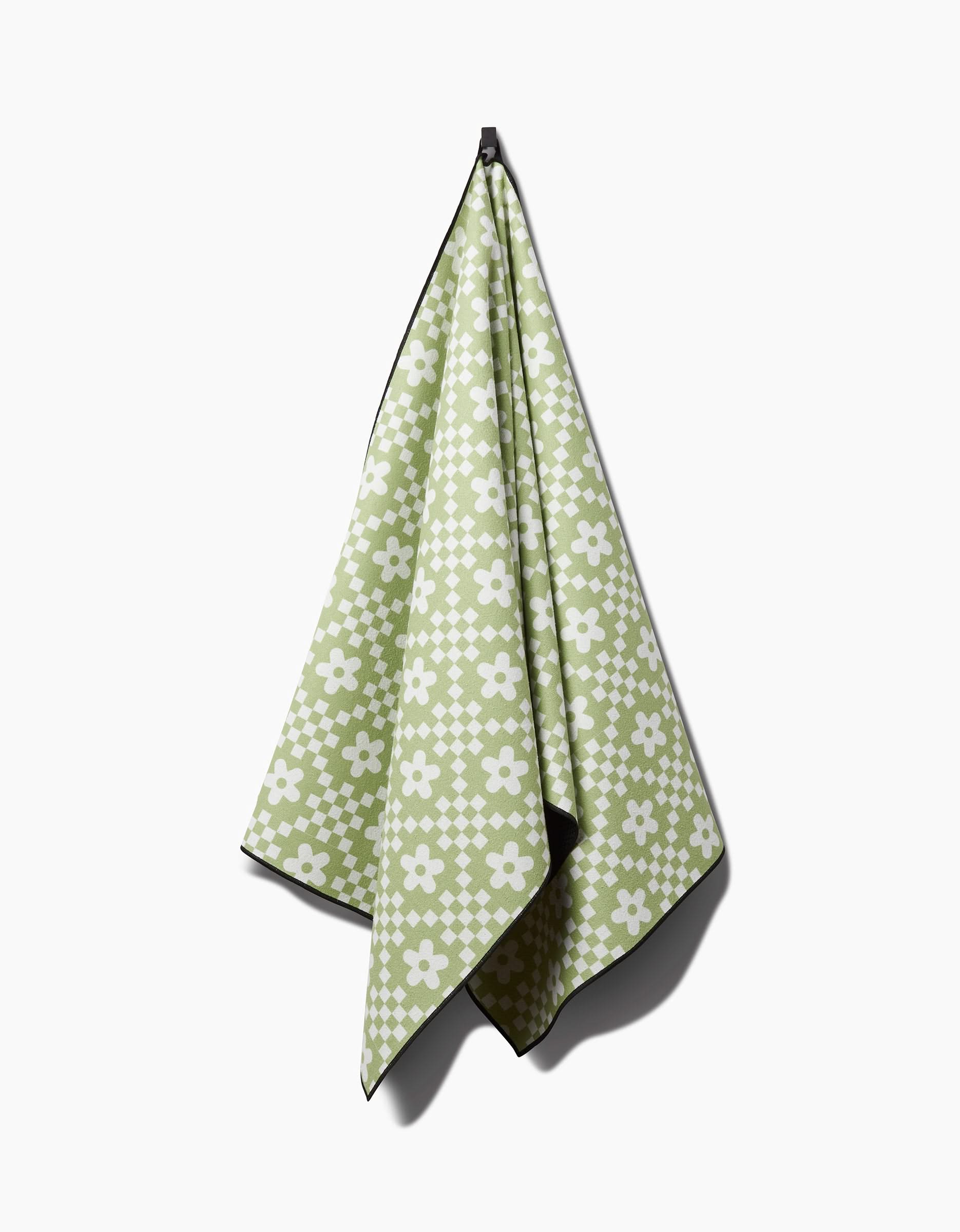 The GEOMETRY Floral Check yoga towel in green, with a white floral and checkerboard design, is shown slip-resistant and hanging on a hook against a plain white background.