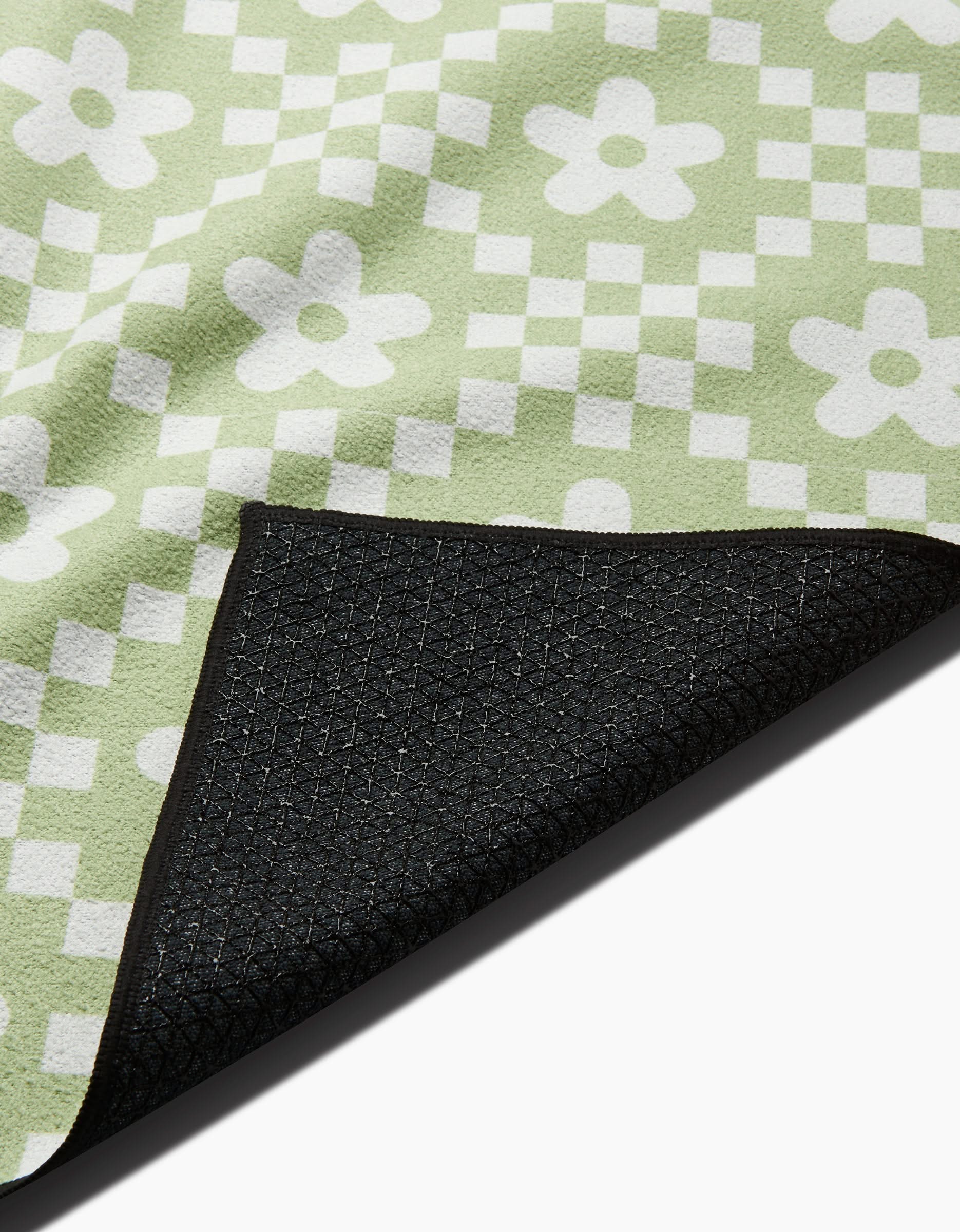 A close-up of the GEOMETRY Floral Check mat shows its green and white checkerboard and floral pattern, with a flipped corner displaying the black, slip-resistant yoga towel underside.
