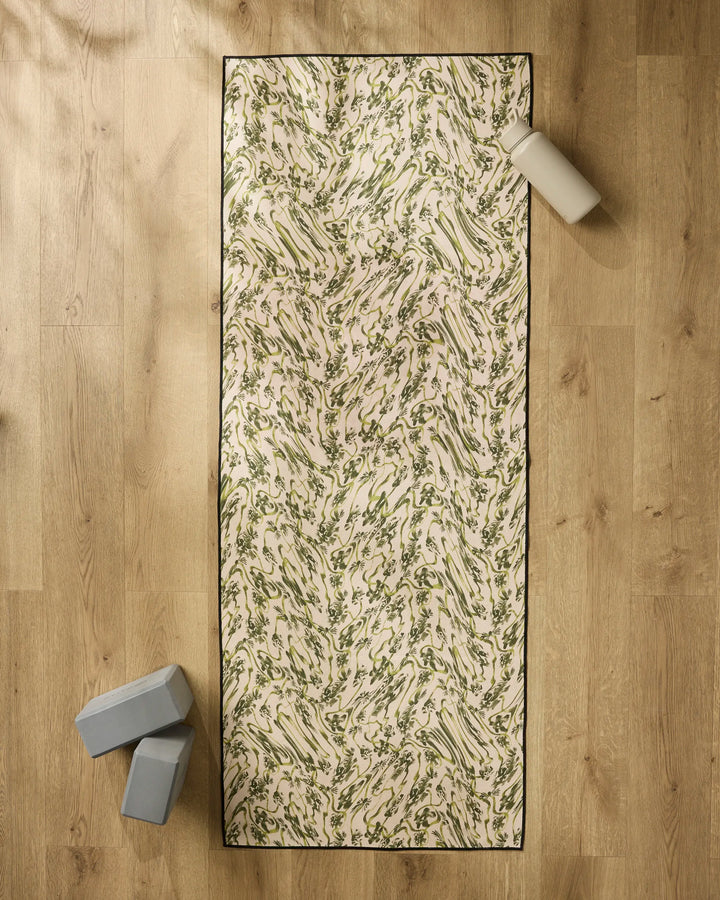 The Geometry Floral Swirls Green yoga mat is laid on a wooden floor with a non-slip towel, two gray yoga blocks at the bottom left, and a white water bottle near the top right corner.