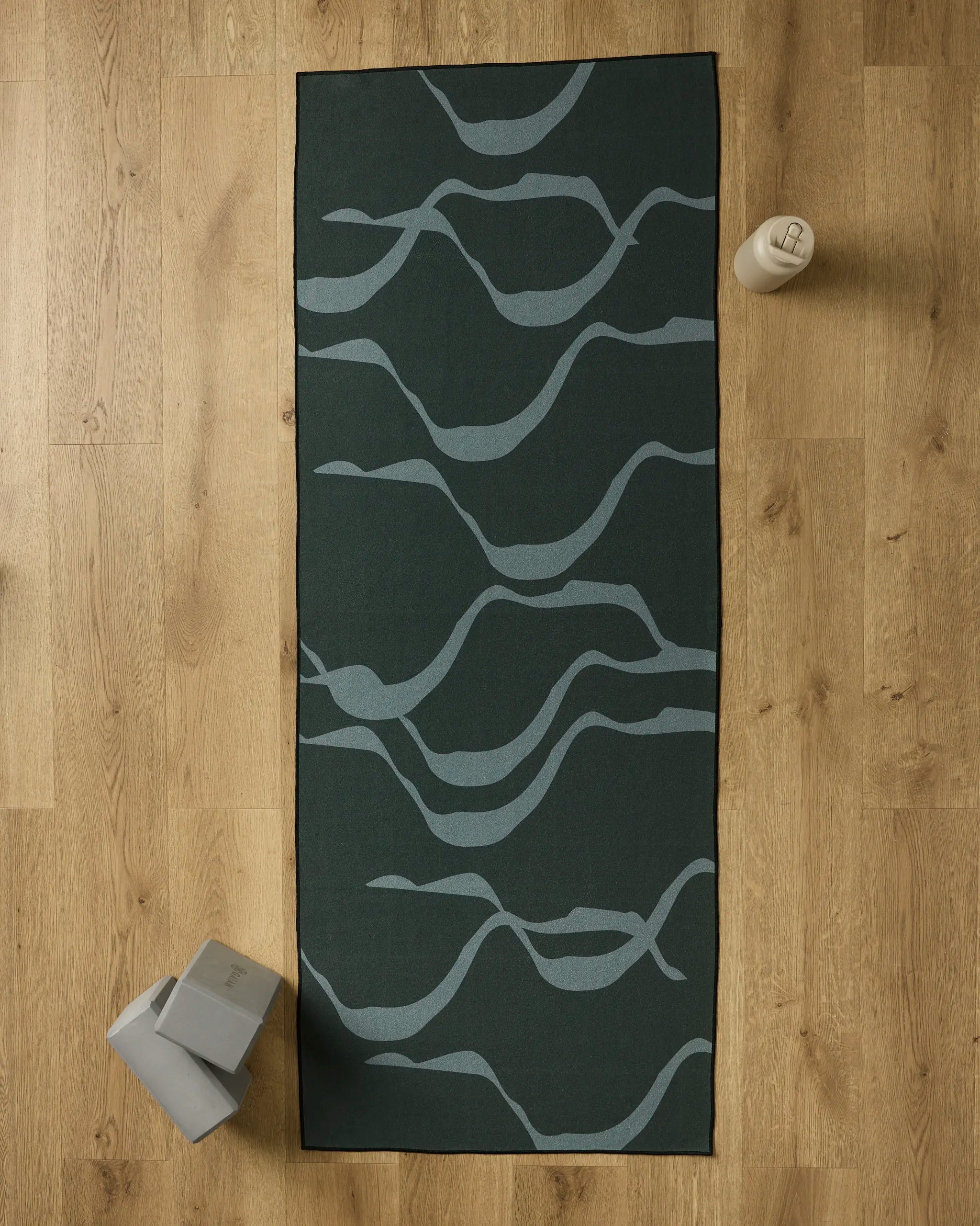 A GEOMETRY Flow yoga mat in dark green with light gray wavy lines lies on a wooden floor. Nearby are a recycled yoga towel, two gray yoga blocks in the bottom left, and a beige water bottle at the top right of the mat.