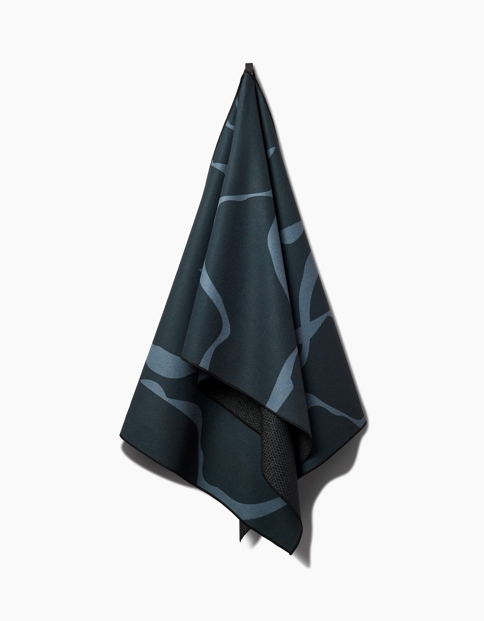 The GEOMETRY Flow yoga towel, made from recycled materials, features an abstract light blue wavy line design on dark fabric and hangs on a hook against a plain white background.