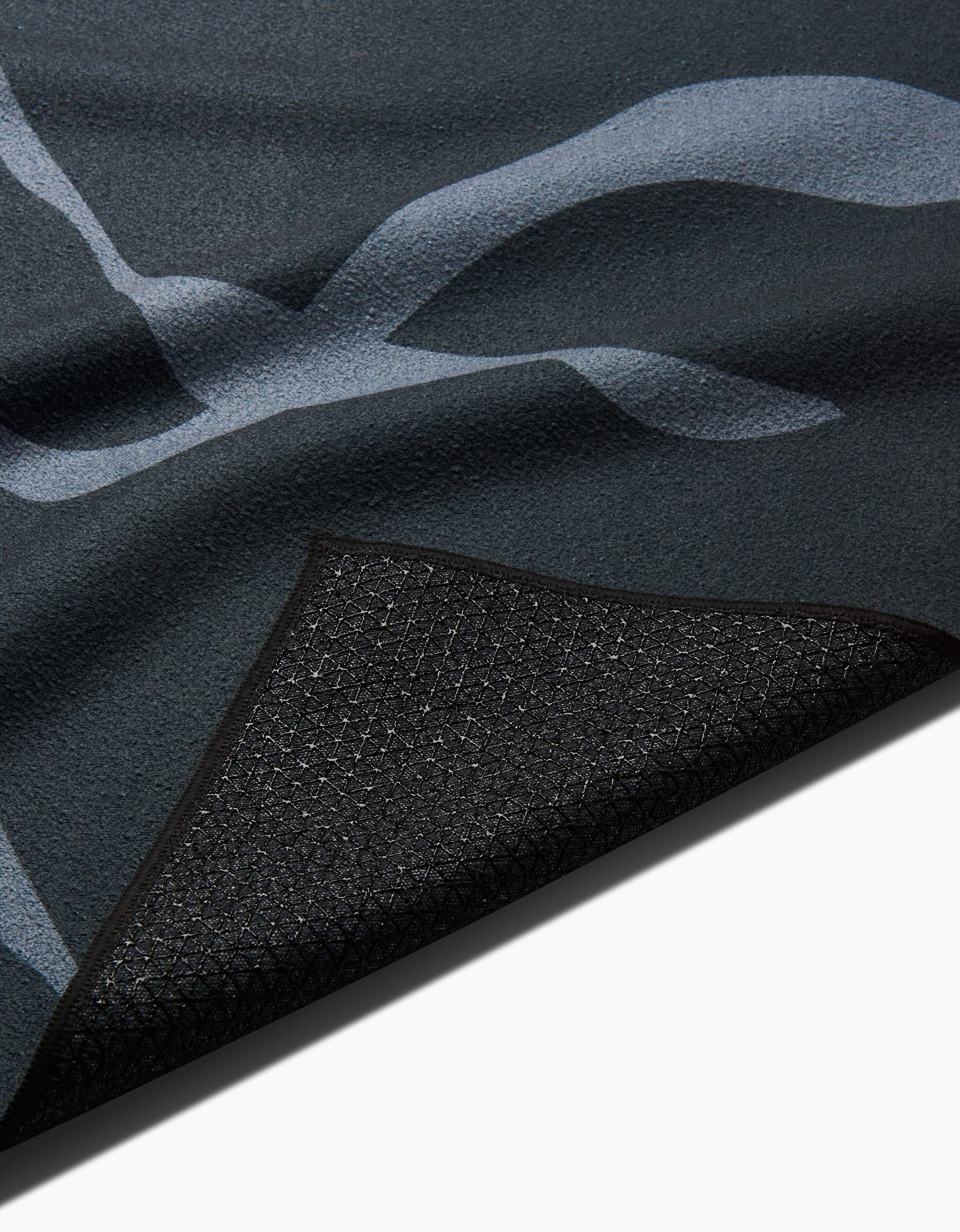 A close-up of GEOMETRY’s Flow mat shows its abstract light blue patterns on a dark surface, with one corner lifted to reveal the textured, non-slip silicone grip backing.