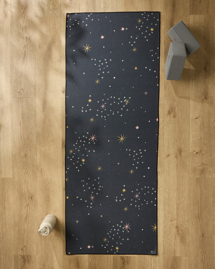 The Ash Cascade Galaxy Burst black yoga mat, featuring a star and dot pattern, rests on a wooden floor with two gray yoga blocks, a white water bottle, and a non-slip towel for extra grip.