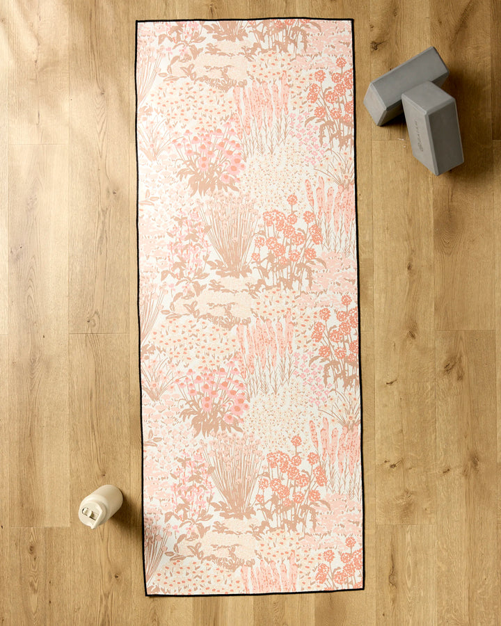 The Garden Bloom yoga mat by Hufton Studio is laid on a wooden floor with a silicone grip towel. A white water bottle rests at the bottom left, and two gray yoga blocks are stacked at the top right.