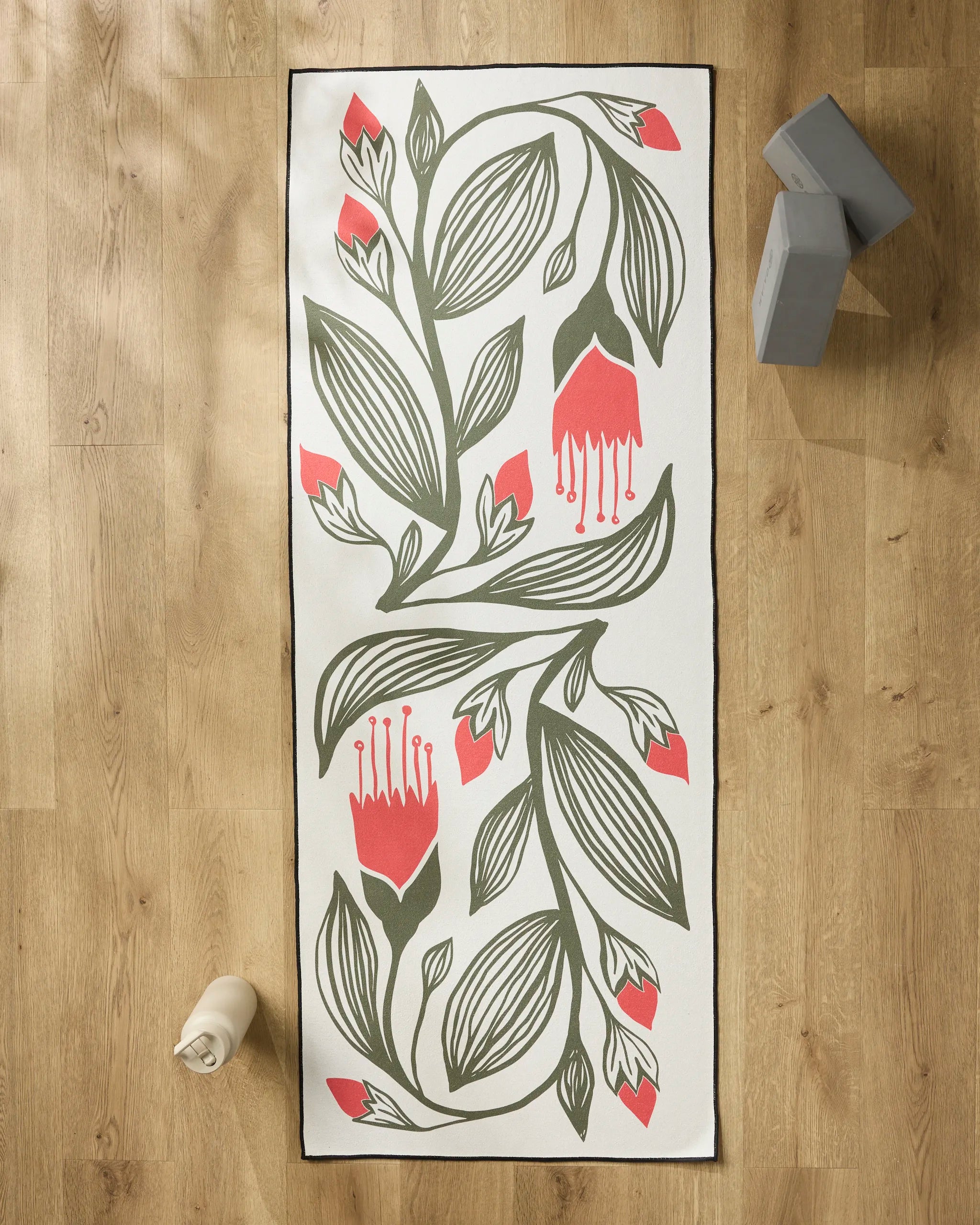 The Ash Cascade Garden Stamp yoga mat, featuring a floral green leaf and red flower design on white, lies on a wooden floor with a water bottle, two gray yoga blocks, and a non-slip towel for enhanced comfort during practice.