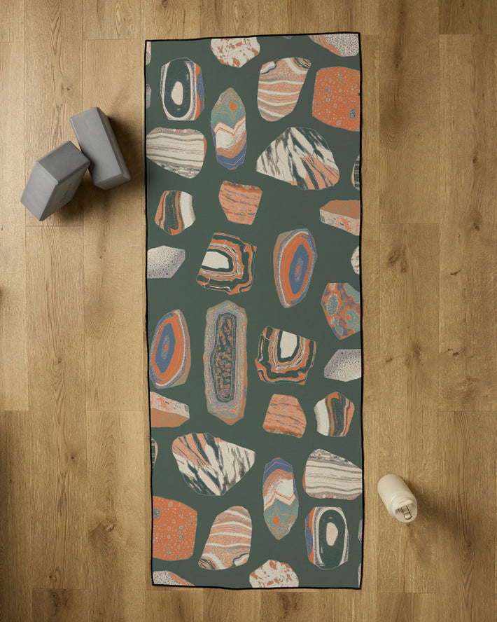 The Geometry Geo Class yoga mat, featuring a colorful abstract rock pattern, rests on a wooden floor with two gray blocks, a white water bottle, and a non-slip towel for enhanced grip during practice.