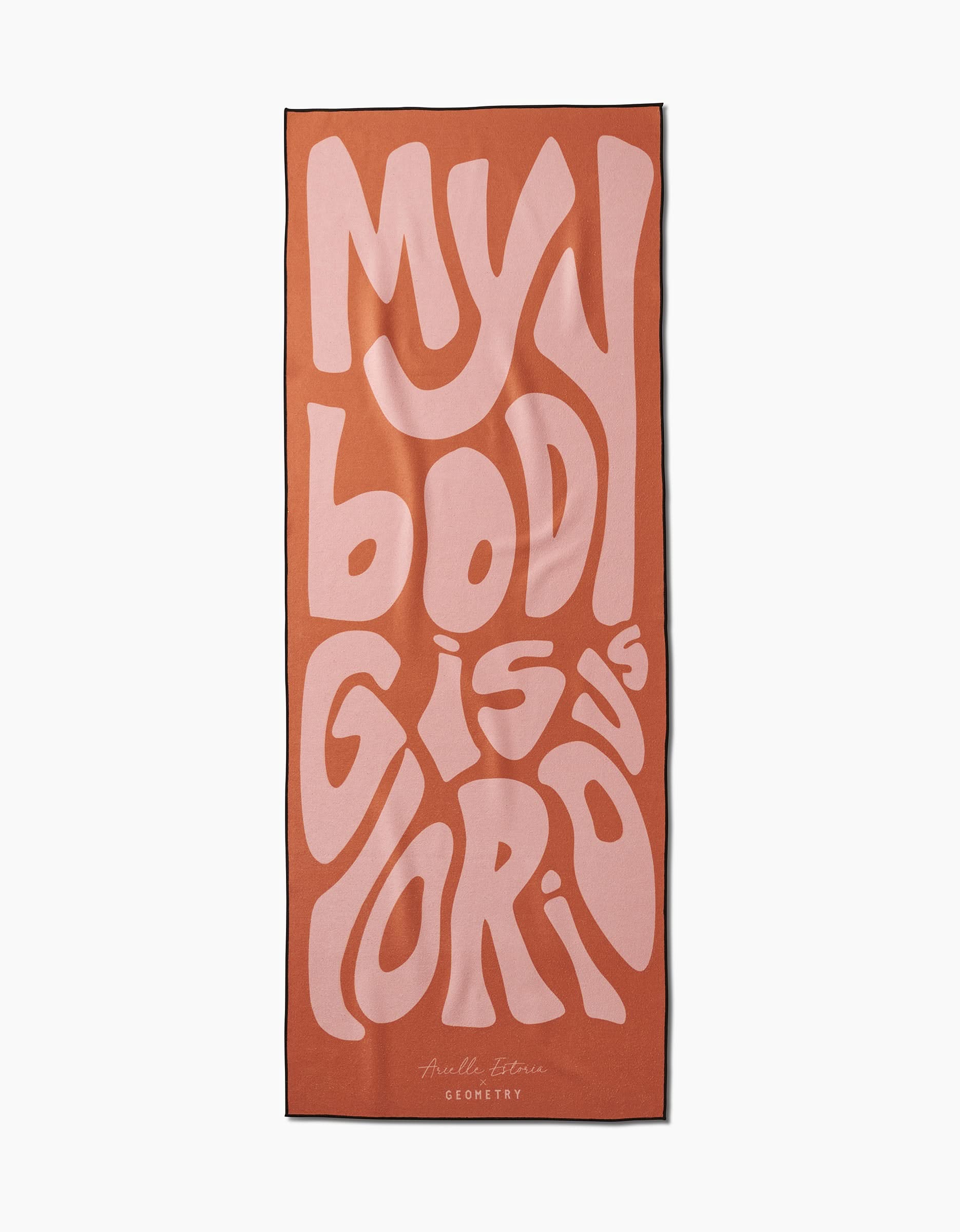 The GLORIOUS yoga towel by Arielle Estoria is rectangular, in orange and pink, with bold wavy MY BODY IS GLORIOUS text and a signature plus GEOMETRY at the bottom. Its non-slip design is perfect for your practice.