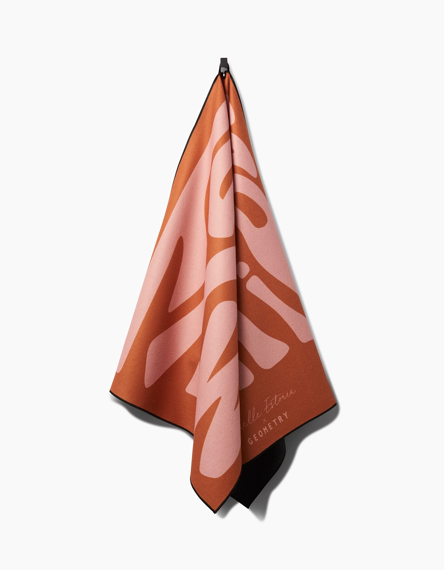 A rust-colored non-slip yoga towel from Arielle Estoria, featuring bold abstract light pink GLORIOUS lettering, hangs from a black hook on a white background. Cursive text appears near the bottom corner.