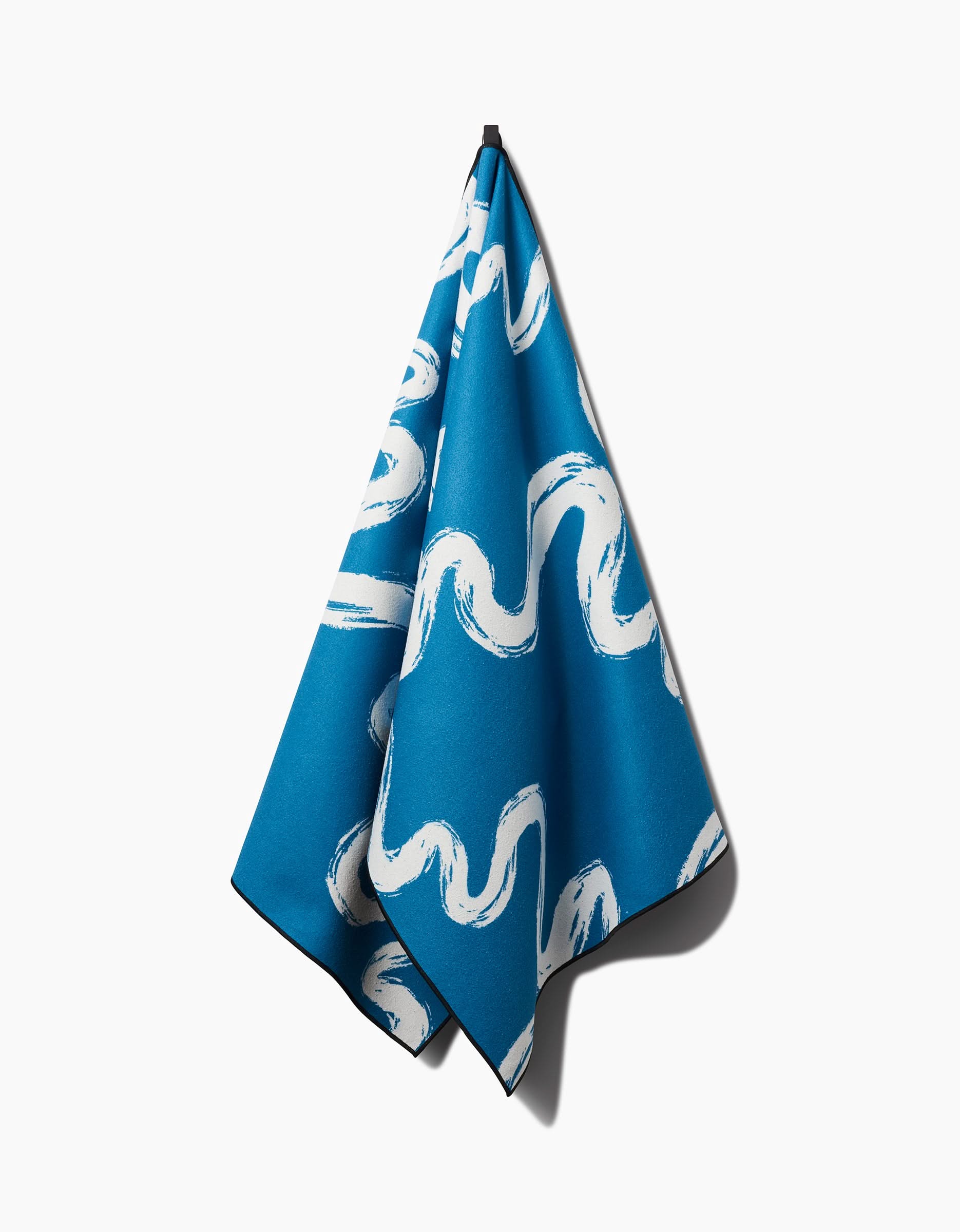 The Elena Fay Go With the Flow blue, slip-resistant yoga towel featuring white wavy brushstroke patterns hangs from a hook against a plain white background.