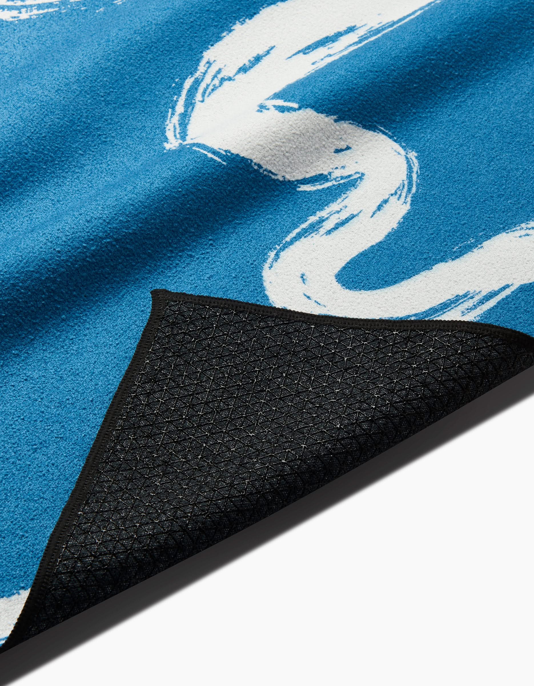 A close-up of Elena Fays Go With the Flow yoga towel, featuring blue fabric with white swirl patterns and a black slip-resistant textured backing, one corner folded to show the underside.