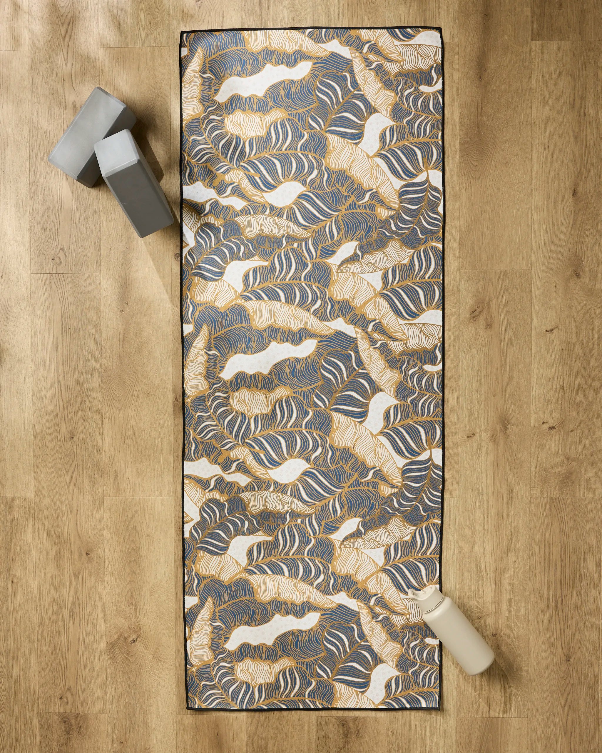 The Golden Palms yoga mat by Marina Solodka, featuring abstract blue, gold, and white designs, lies on a wooden floor. A non-slip towel covers the mat with two gray yoga blocks and a light water bottle placed beside it.