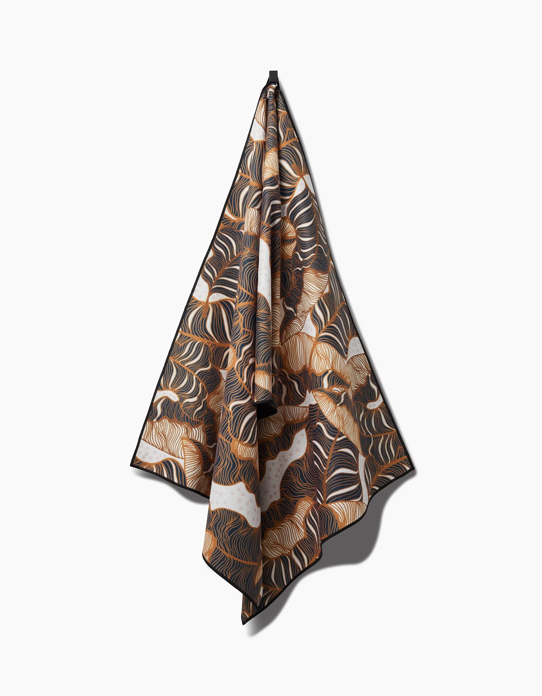The Marina Solodka Golden Palms yoga towel, featuring a patterned slip-resistant design in swirling brown, black, and white abstract motifs, hangs neatly from a single point against a plain white background with a soft shadow.