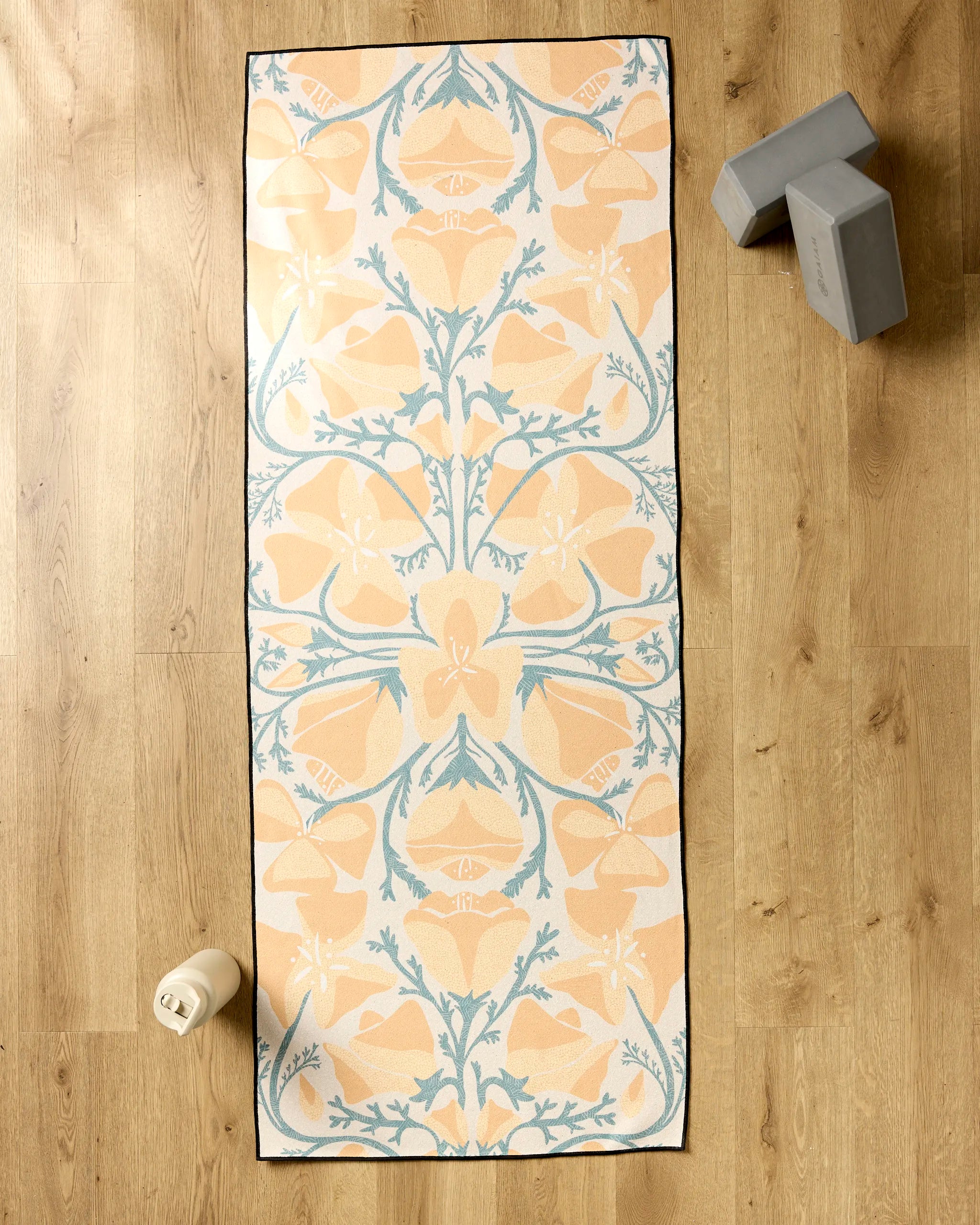 The Golden Poppy yoga mat by Julianne Haness, featuring a beige and blue floral pattern, is laid out on a wooden floor with two gray blocks, a silicone grip towel, and a white water bottle nearby.
