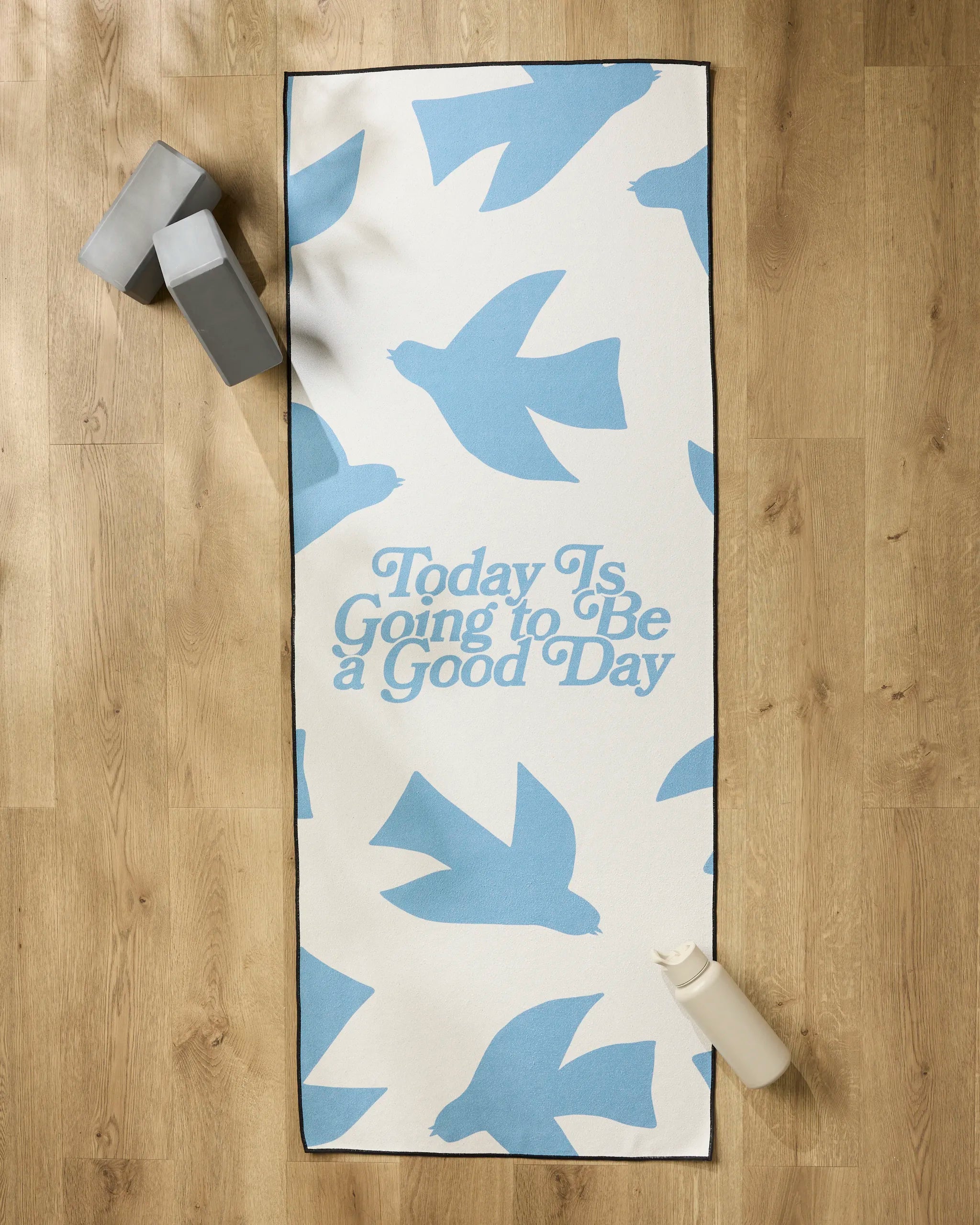 The Quotes by Christie "Good Day" non-slip yoga towel featuring blue bird silhouettes and the phrase "Today Is Going to Be a Good Day" is shown on a wooden floor with two yoga blocks and a water bottle nearby.