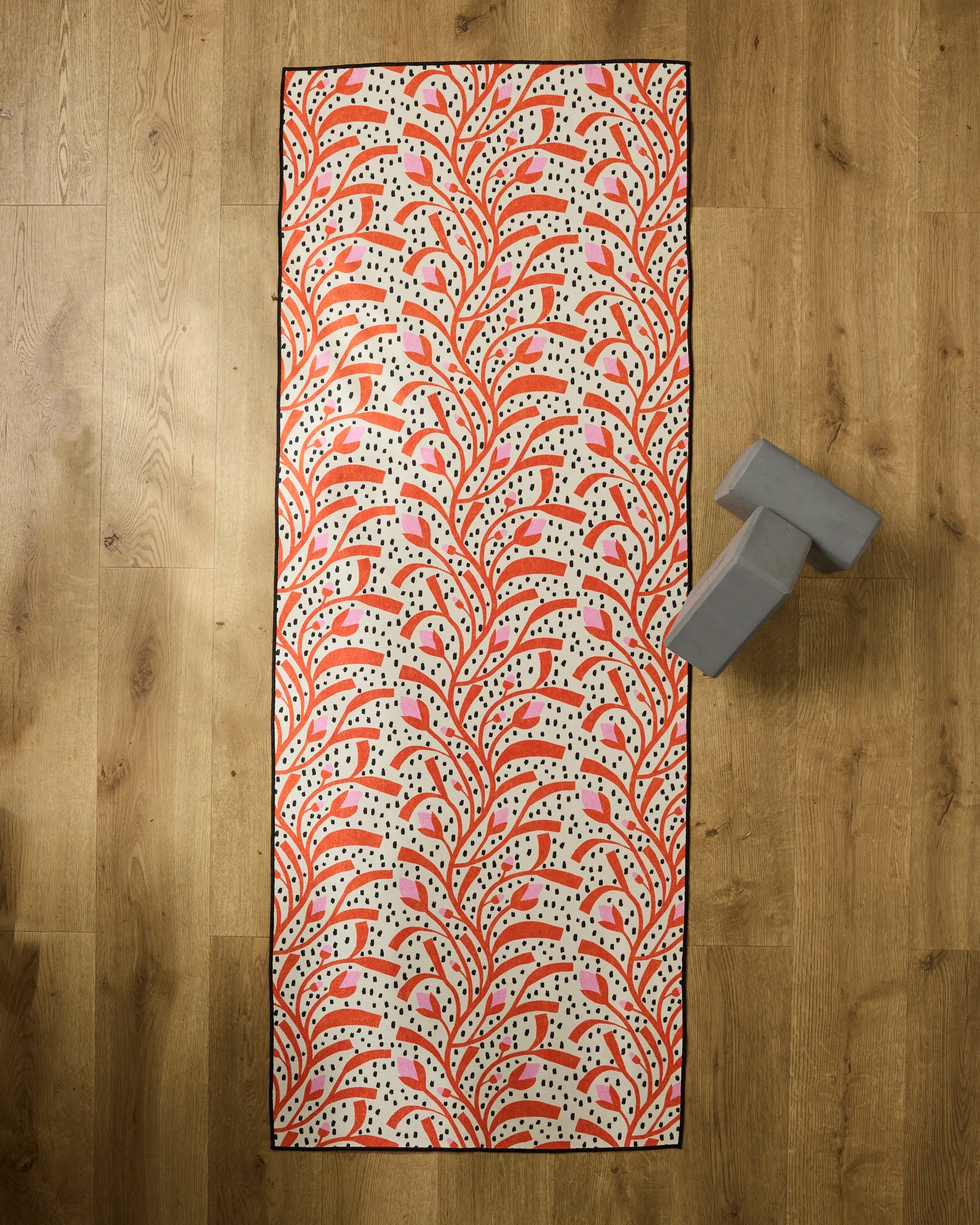 The Claire Iglesias "Graphic Flower Buds" rectangular yoga mat, featuring a red and pink leafy pattern on cream, is placed on a wooden floor with a silicone grip towel and a gray yoga block near the top right corner.