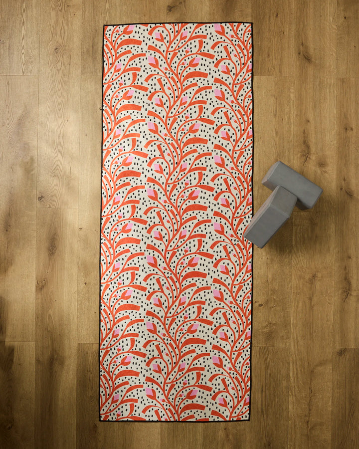 The Claire Iglesias "Graphic Flower Buds" rectangular yoga mat, featuring a red and pink leafy pattern on cream, is placed on a wooden floor with a silicone grip towel and a gray yoga block near the top right corner.