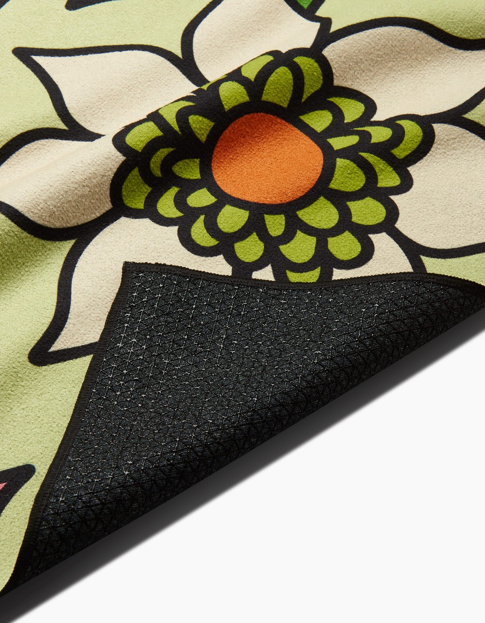 A close-up of GEOMETRYs Green Flowers yoga towel with white petals, green segments, and an orange center. The flipped corner shows a textured black underside crafted from recycled materials.