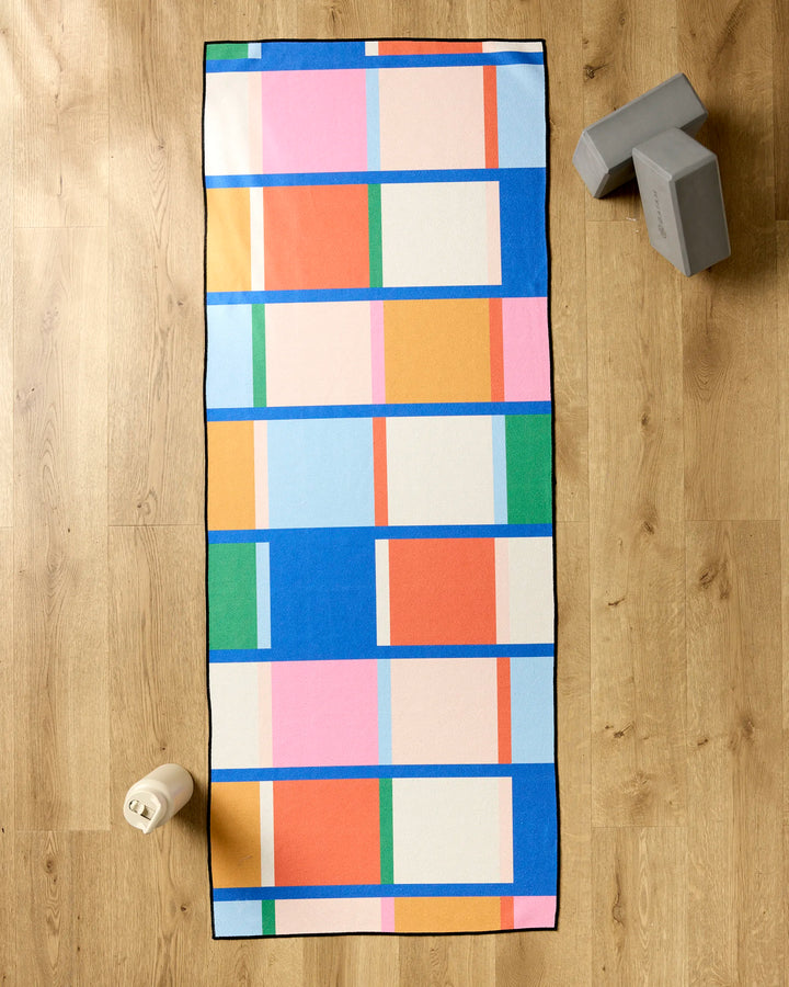The Grid yoga mat by Jennifer Bouron features a colorful geometric rectangle pattern and rests on a wooden floor alongside two gray yoga blocks, a white water bottle, and a recycled yoga towel.