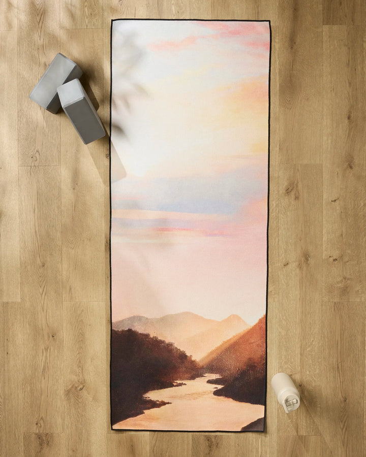 The Her Majesty yoga mat by Stefanie Bales, featuring a sunset mountain landscape, is displayed on a wooden floor with two gray yoga blocks, a white water bottle, and a non-slip yoga towel for extra stability during your practice.