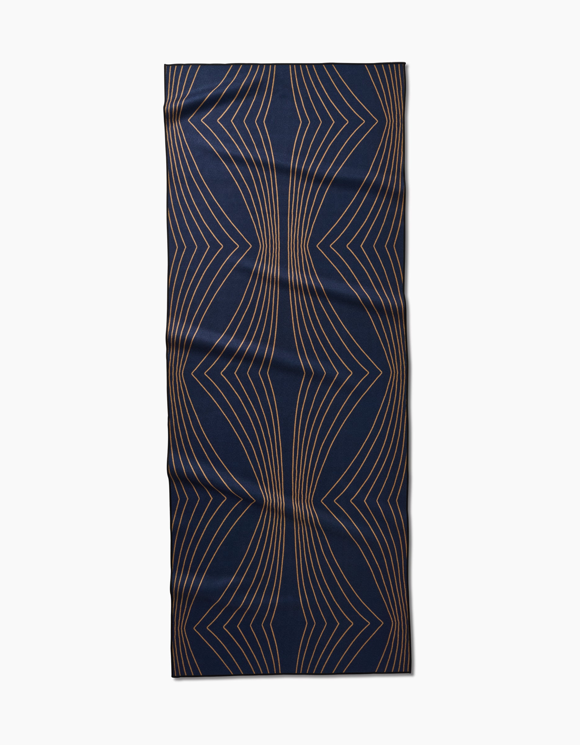 The Hera yoga towel by Amy MacCready features a rectangular, dark blue non-slip surface with curved, symmetrical gold geometric lines in a wavy pattern. It is shown lying flat on a white background.