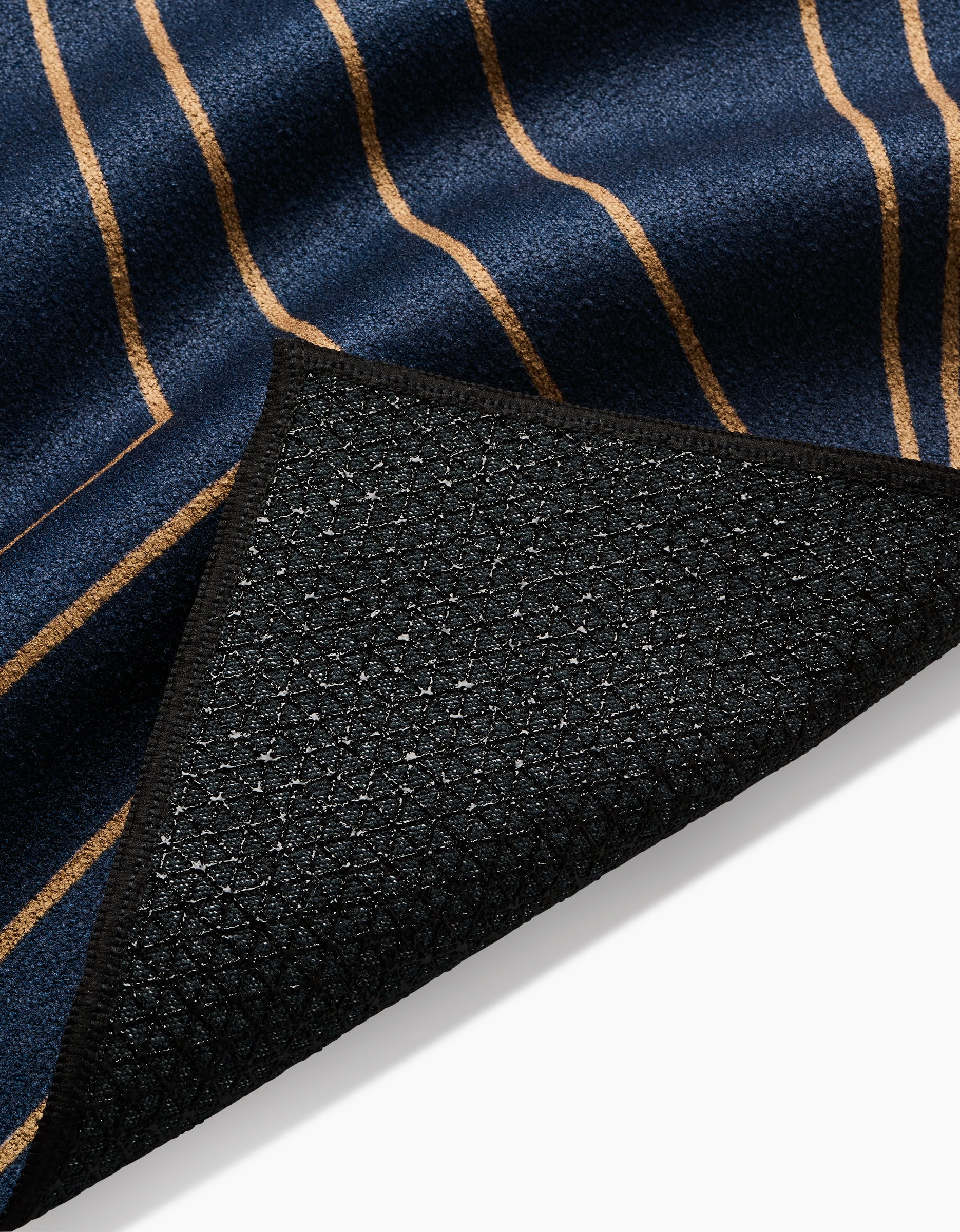 A close-up of Amy MacCready’s Hera yoga towel shows its navy blue fabric with thin tan stripes and a textured black, non-slip recycled underside. A folded corner reveals the contrast between the front and grippy backing.