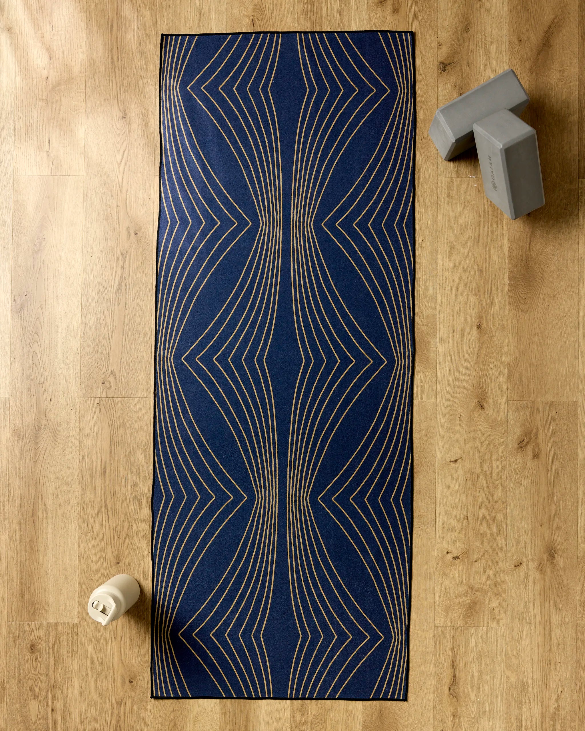 The Hera yoga mat by Amy MacCready, featuring blue with beige geometric lines, is shown on a wooden floor with a white water bottle, two gray yoga blocks, and a non-slip yoga towel for extra grip during your practice.