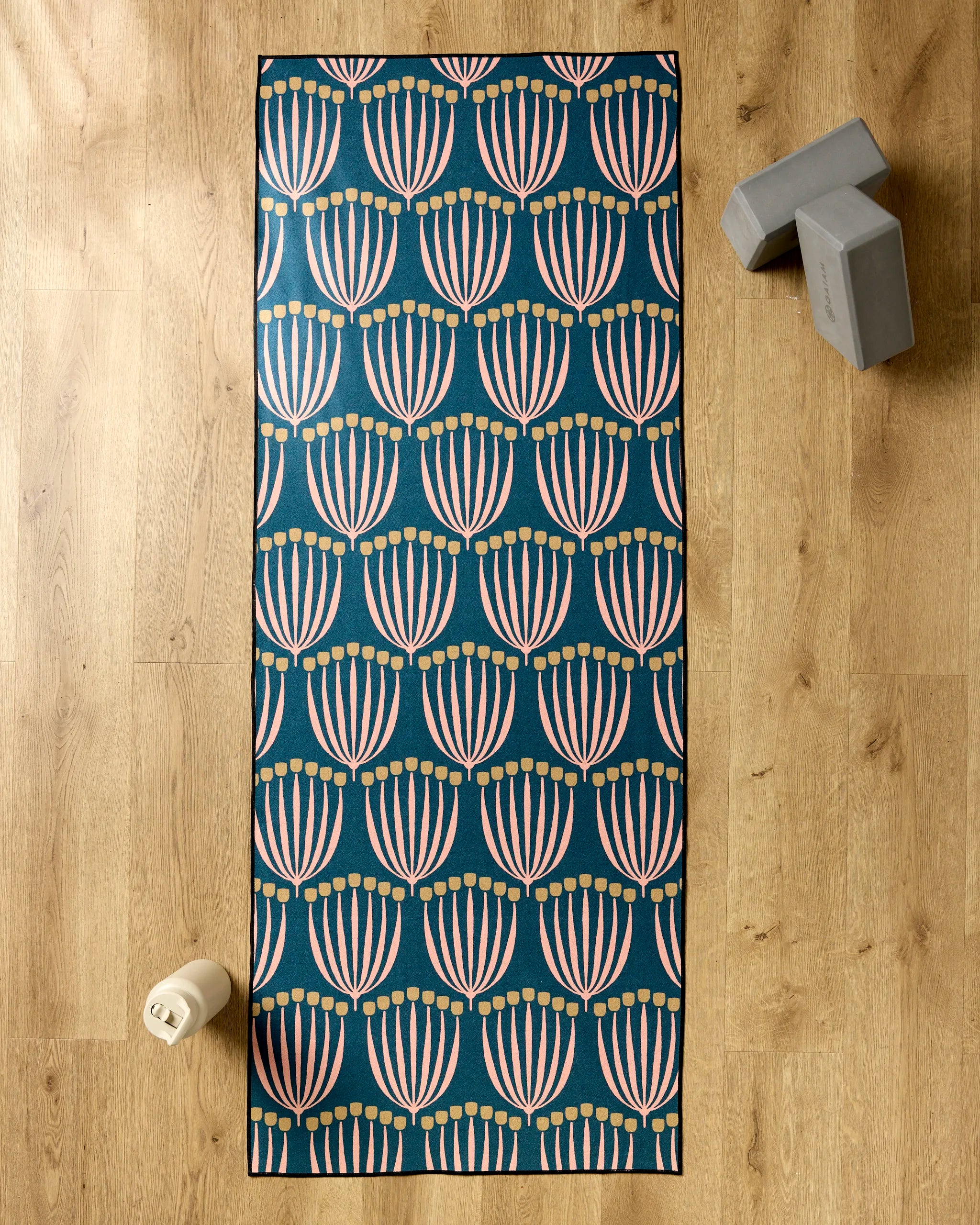 The Josie yoga mat by Amy MacCready, featuring pink and yellow floral patterns on teal, is displayed on a wooden floor with a non-slip yoga towel, two gray yoga blocks, and a white water bottle.