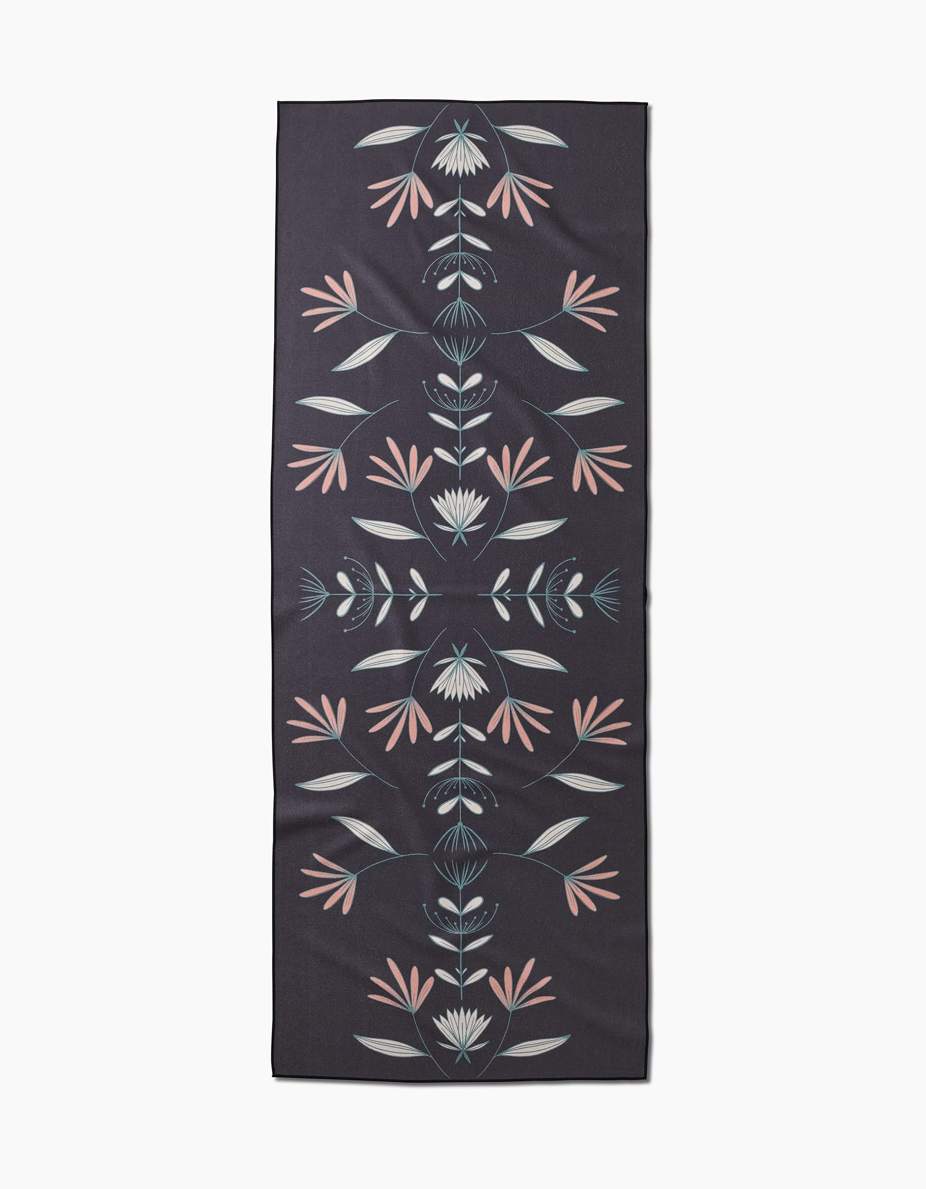 The Jungle Maze yoga mat by Amita Nair is a rectangular, dark-hued mat made from recycled materials, featuring a symmetrical floral pattern with stylized leaves and flowers in muted pink, blue, and gray.