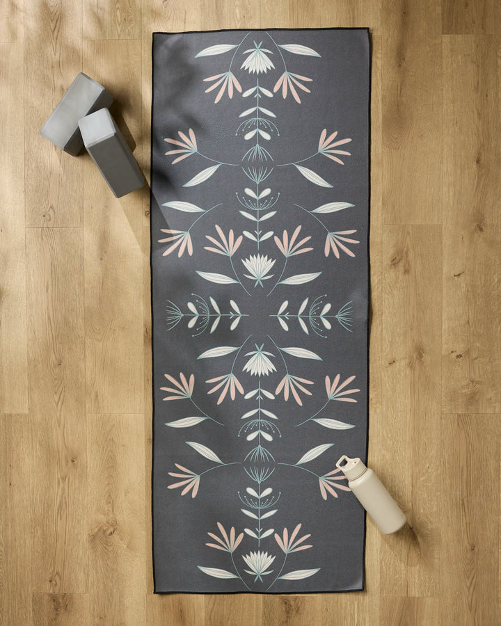 The Amita Nair Jungle Maze yoga mat, featuring a dark gray base with a pastel floral design, is spread on a wooden floor. A slip-resistant yoga towel covers it, with two gray blocks at the top left and a white water bottle at the bottom right.