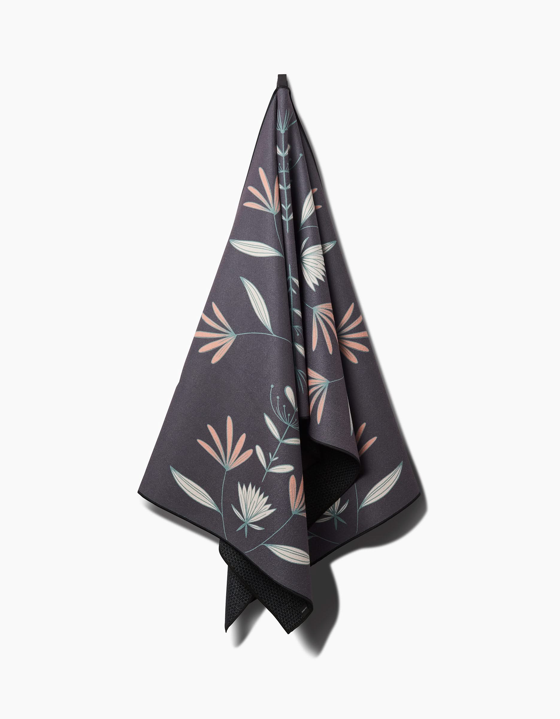 The Amita Nair Jungle Maze yoga towel, in dark tones with a subtle floral pattern in pink, white, and green, hangs on a hook against a white background and features a silicone grip for extra stability.