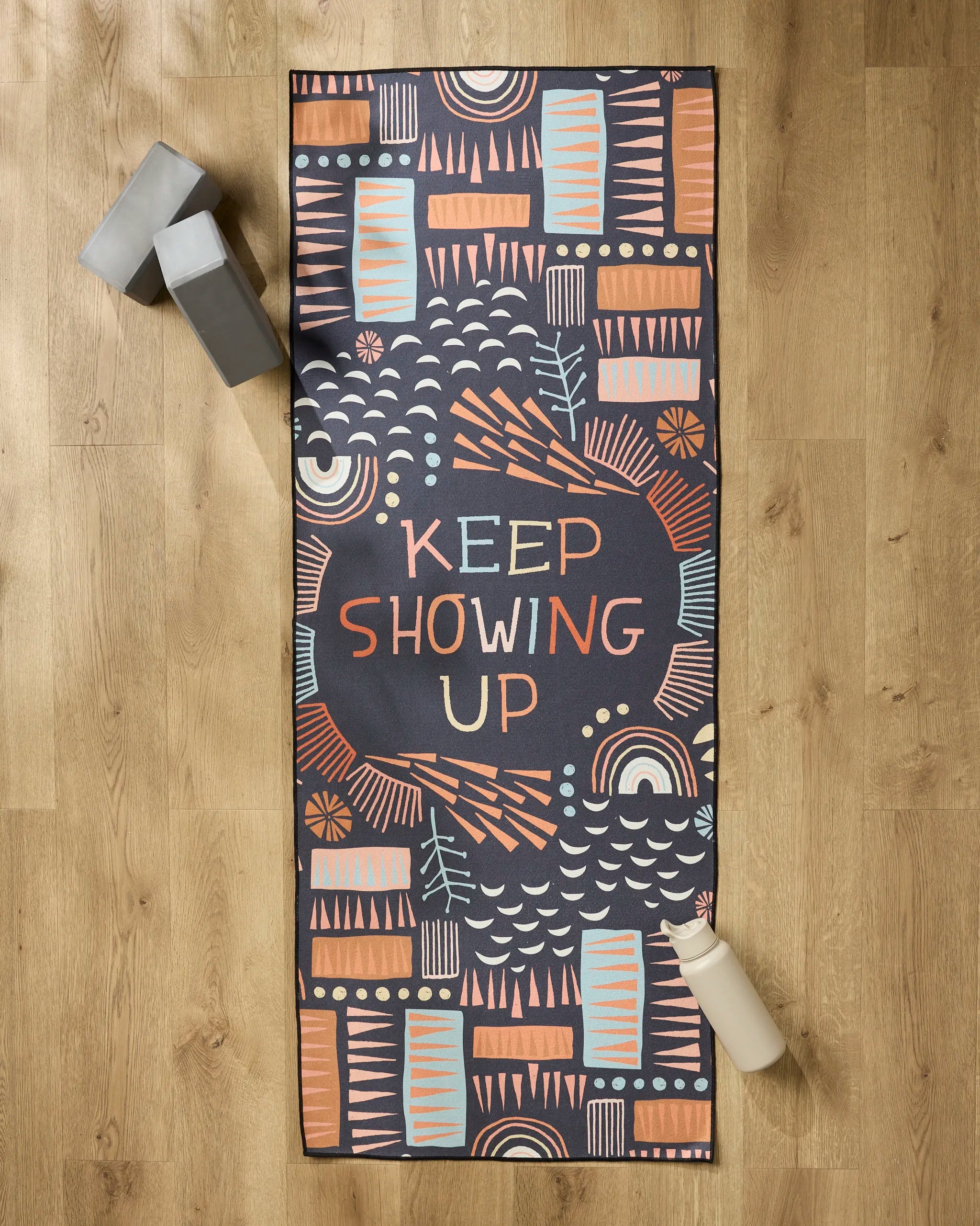 The Jessica Swift "Keep Showing Up" patterned yoga mat is on a wooden floor, accompanied by a recycled yoga towel, two gray yoga blocks, and a white water bottle.