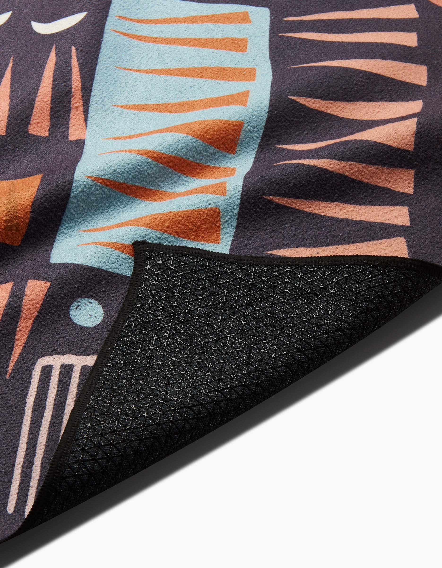 A close-up of the Keep Showing Up yoga towel by Jessica Swift, featuring a geometric pattern in orange, light blue, and peach on dark fabric. A flipped corner reveals the slip-resistant, recycled black underside.
