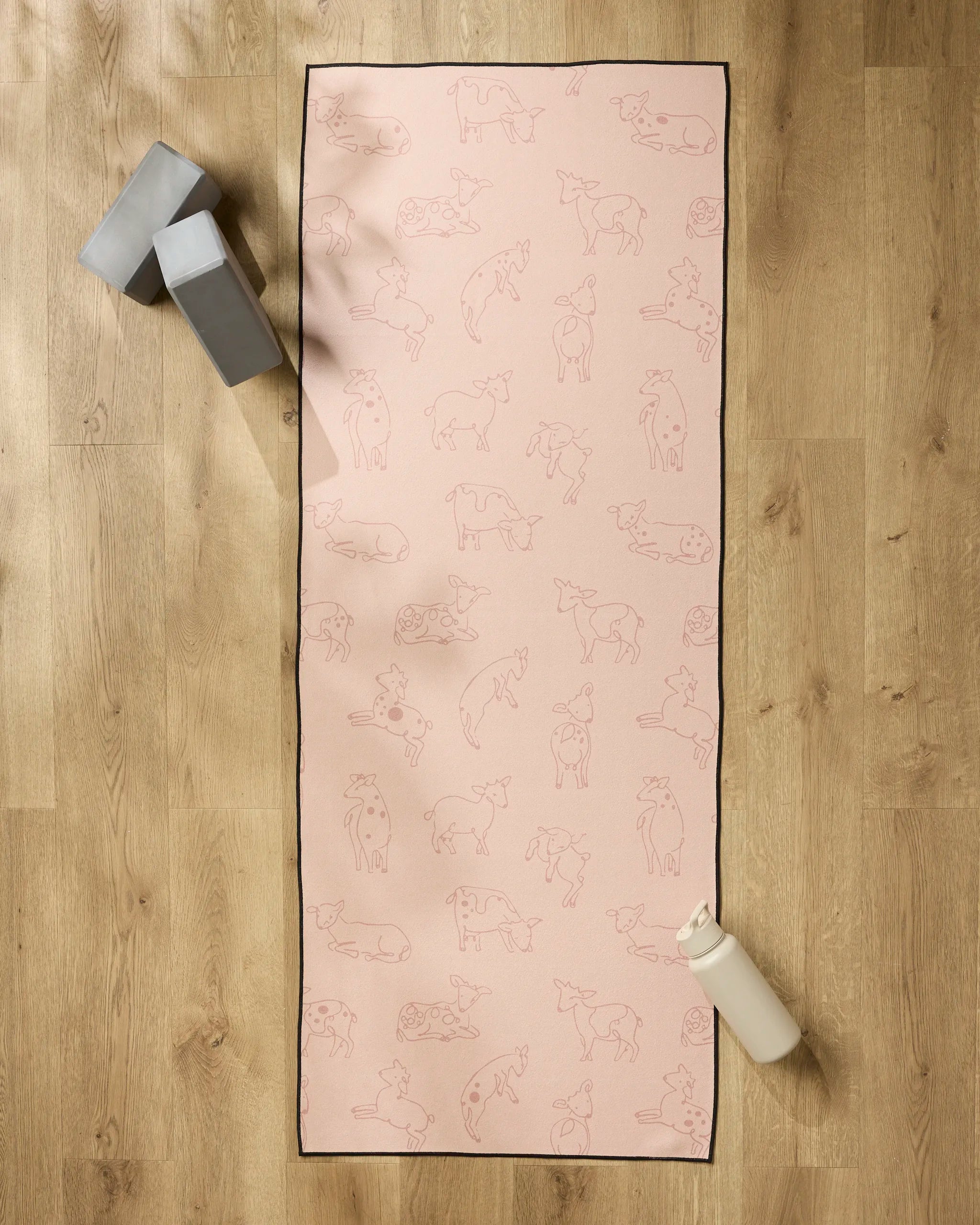 The Sara Mari Kidding Around pink yoga mat with animal line drawings is spread on a wooden floor, topped with a non-slip towel. Two gray yoga blocks and a white water bottle are placed next to the mat.