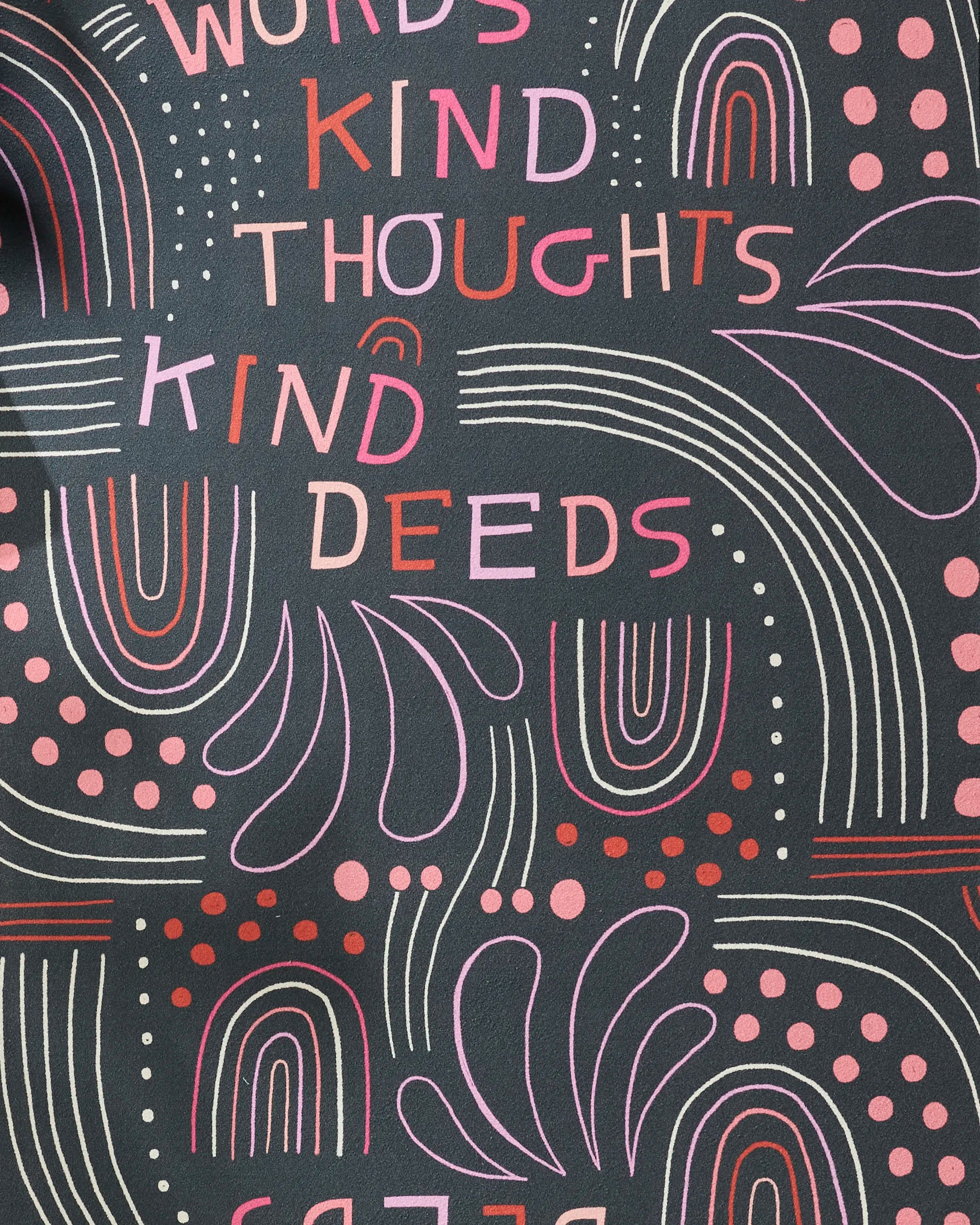 The Jessica Swift "Kind Hearts" yoga towel features a non-slip recycled design with abstract shapes, rainbows, dots, and lines in pink and red on black. “KIND WORDS, KIND THOUGHTS, KIND DEEDS” appears in playful colors across the towel.