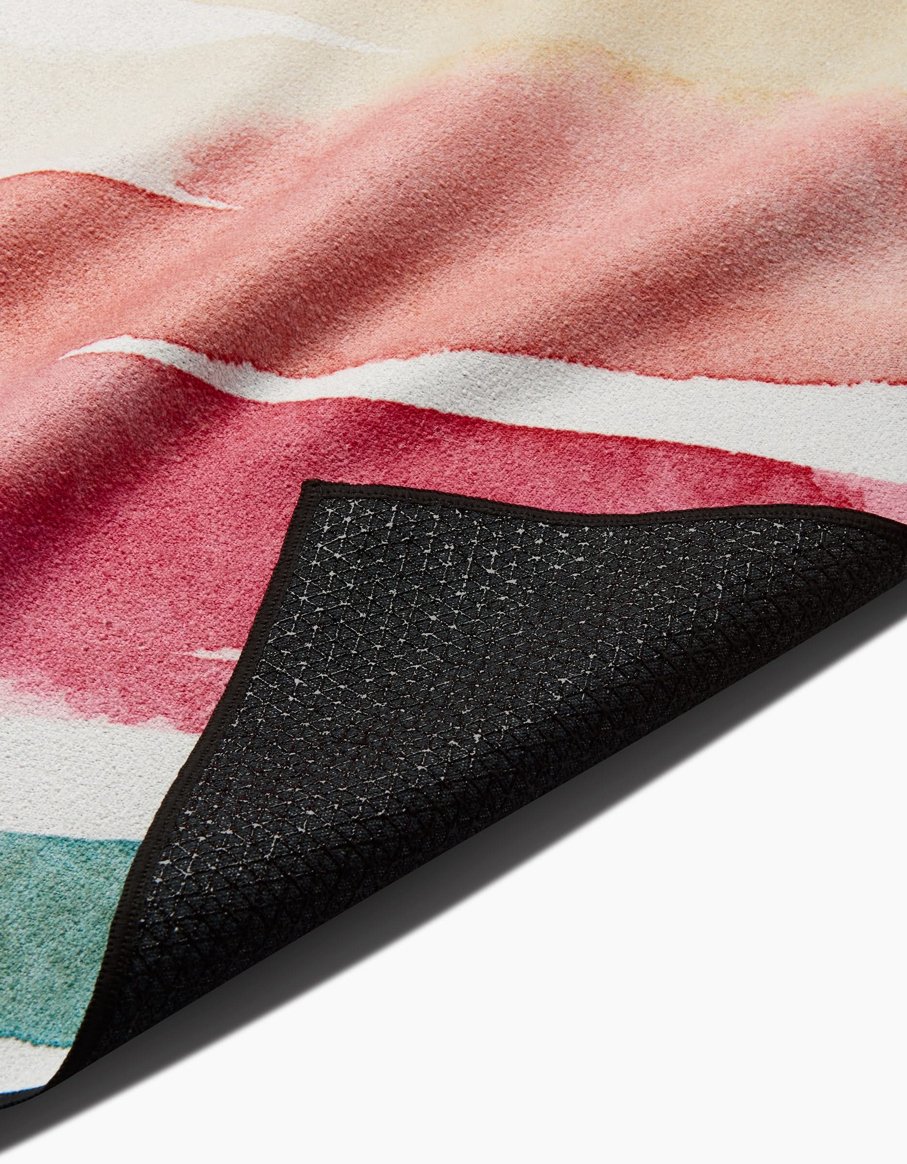 A close-up of the Sara Mari Lakeside area rug shows its vibrant red, pink, and green abstract watercolor patterns. A lifted corner reveals a black textured non-slip backing made from recycled materials.
