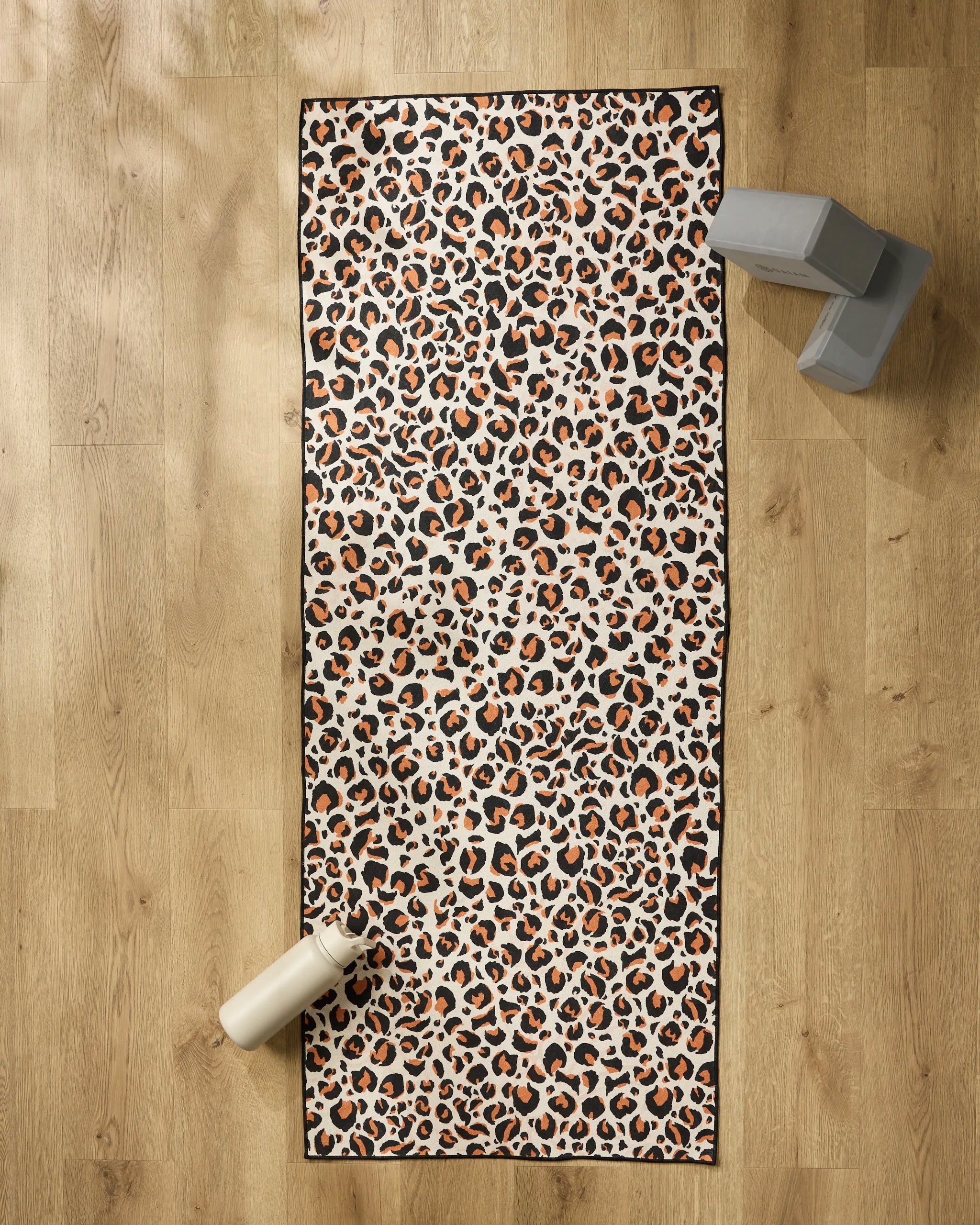 The GEOMETRY Leopard yoga mat is spread on a light wooden floor with a white water bottle at one end, two gray yoga blocks stacked in the top right corner, and a non-slip towel neatly draped alongside.