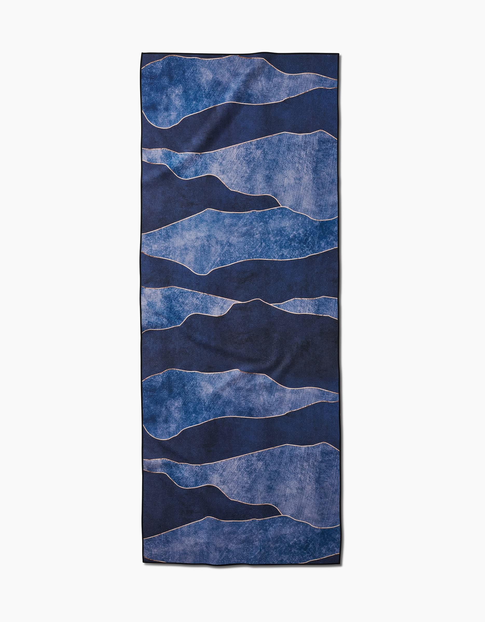 The GEOMETRY Lined With Gold yoga towel is rectangular, made from recycled materials, and features an abstract design with wavy blue bands on a light background.