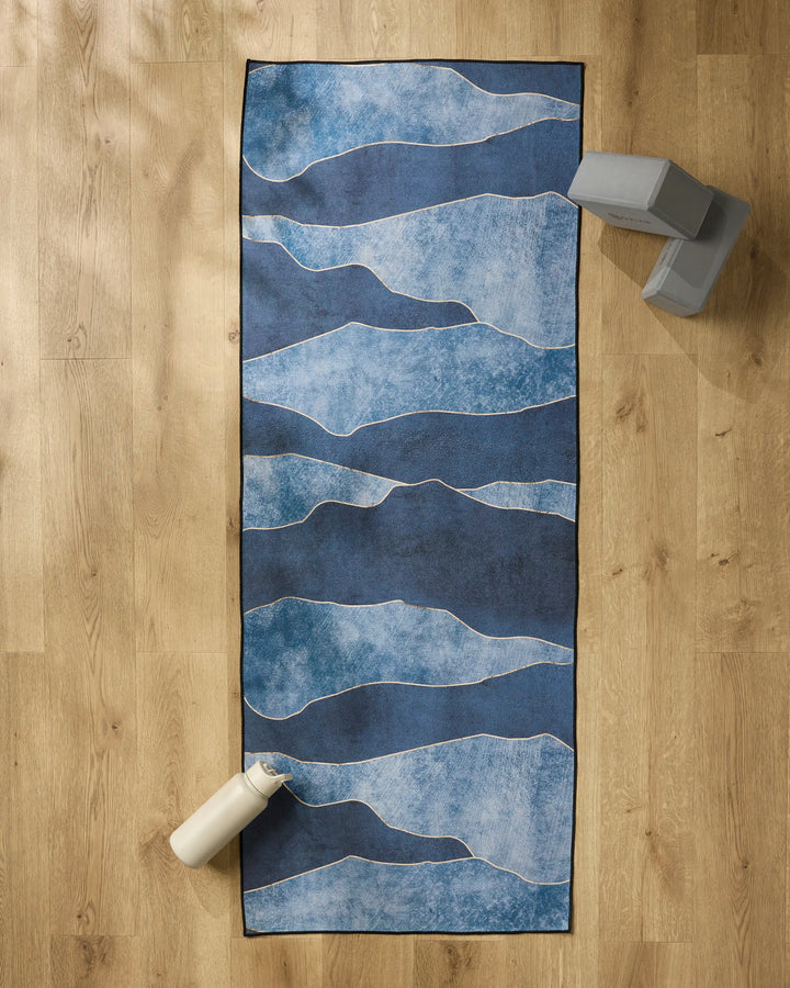 The GEOMETRY "Lined With Gold" yoga mat featuring blue with wavy white lines is spread on a wooden floor, topped with a slip-resistant towel. Two gray blocks are in the top right, and a white water bottle sits at the bottom left.