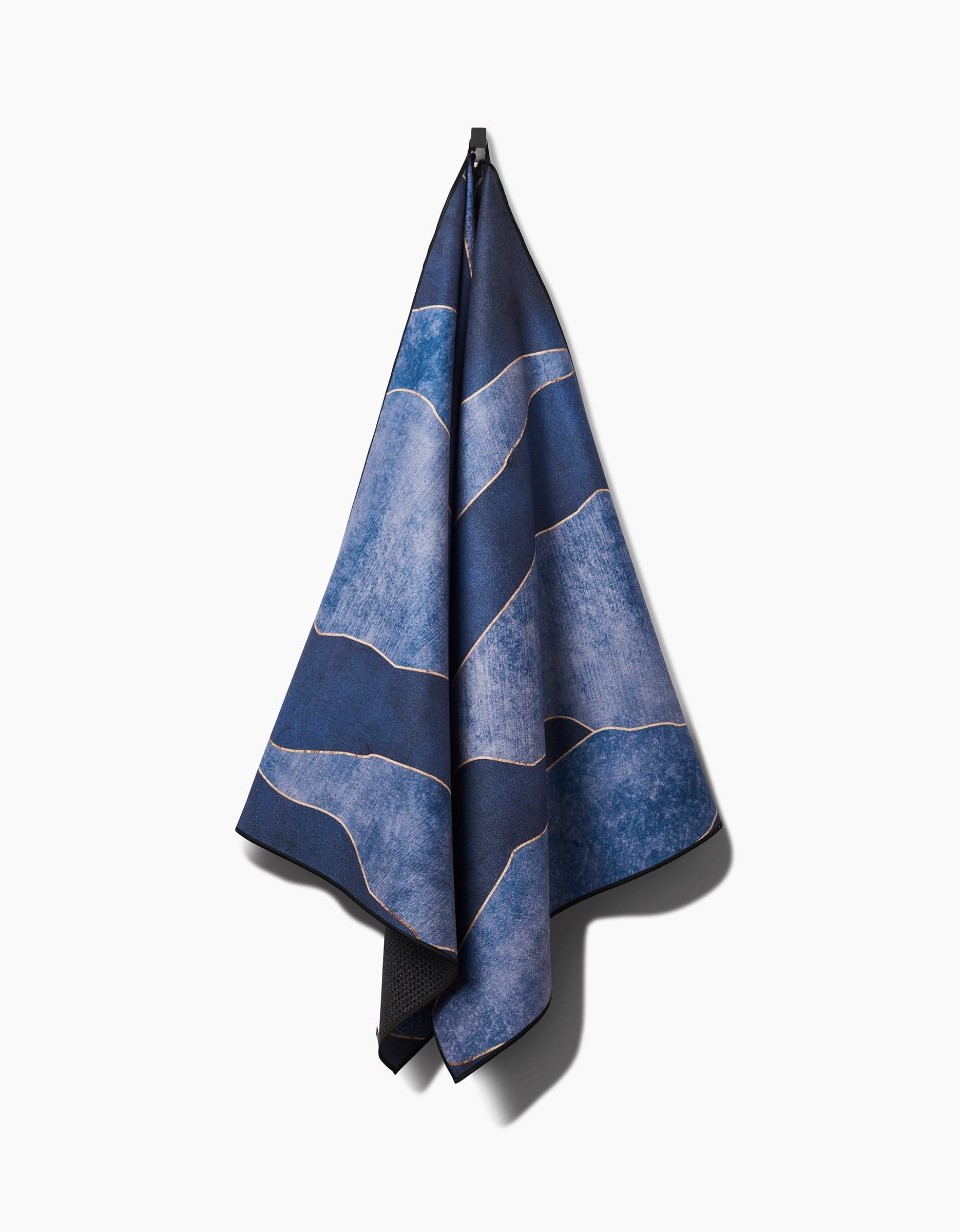 The GEOMETRY Lined With Gold yoga towel, made from recycled materials, features blue tones with light wavy lines and hangs from a hook against a plain white background.