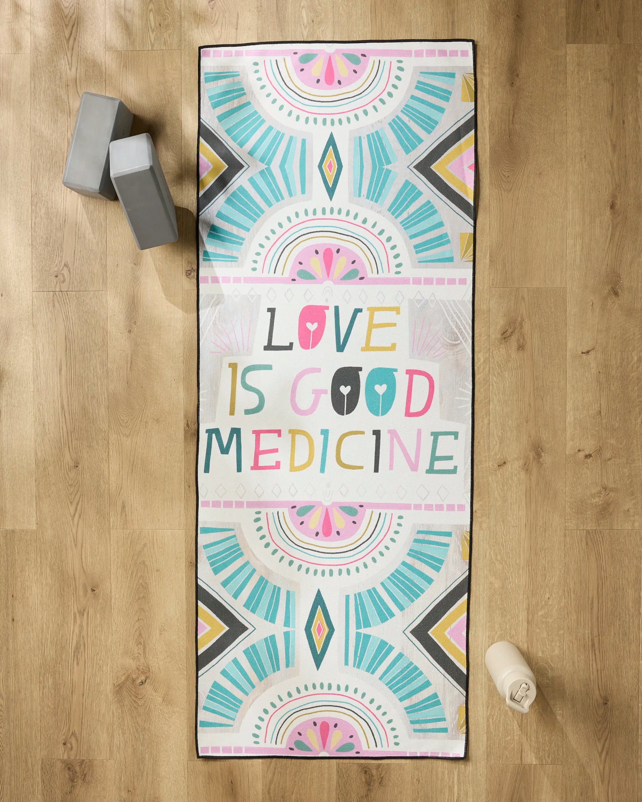 The Love is Good Medicine yoga mat by Jessica Swift features vibrant geometric patterns and the phrase in multicolored letters, accompanied by a non-slip towel, gray yoga blocks, and a water bottle on a wooden floor.
