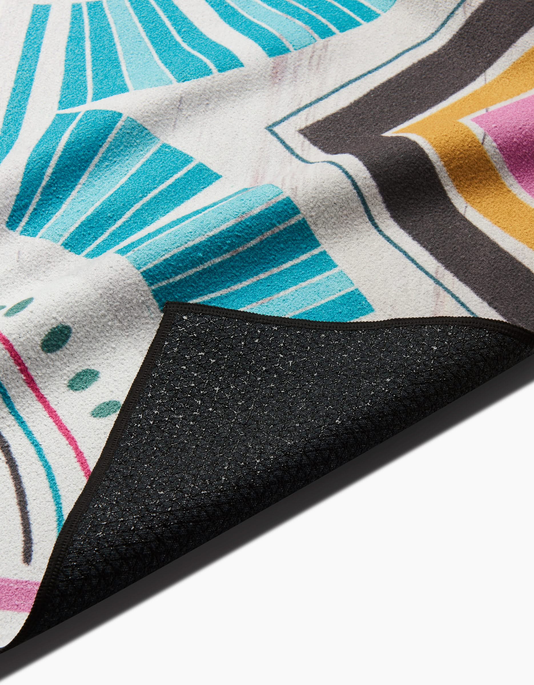 A close-up of the Love is Good Medicine yoga towel by Jessica Swift, featuring a vibrant geometric pattern in blue, yellow, pink, and black with one corner flipped to show its textured, non-slip black underside.